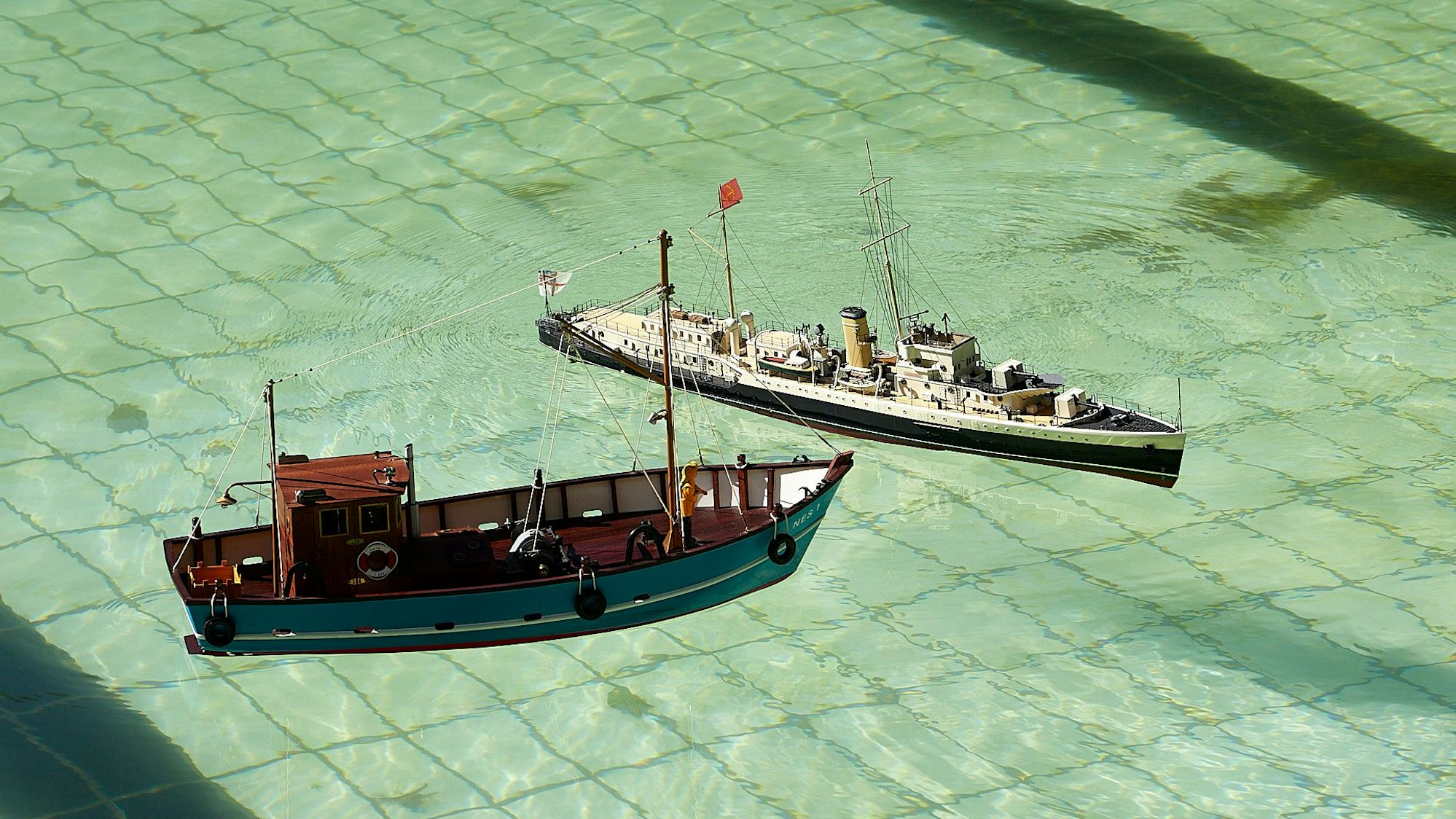 Zwei Modellschiffe sind auf dem Wasser des Freibads, auf dem Grund sind die Kacheln des Schwimmbeckens zu sehen.