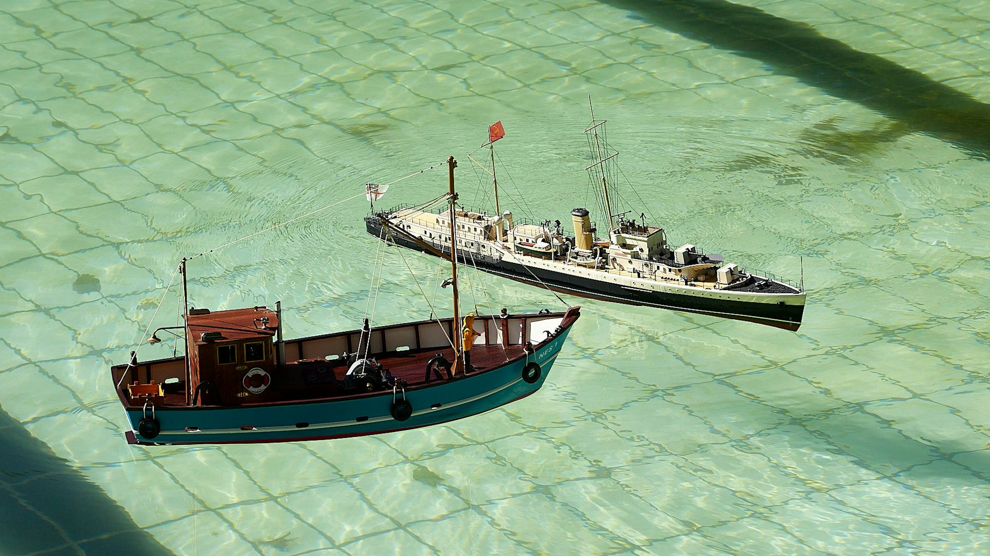 Zwei Modellschiffe sind auf dem Wasser des Freibads, auf dem Grund sind die Kacheln des Schwimmbeckens zu sehen.