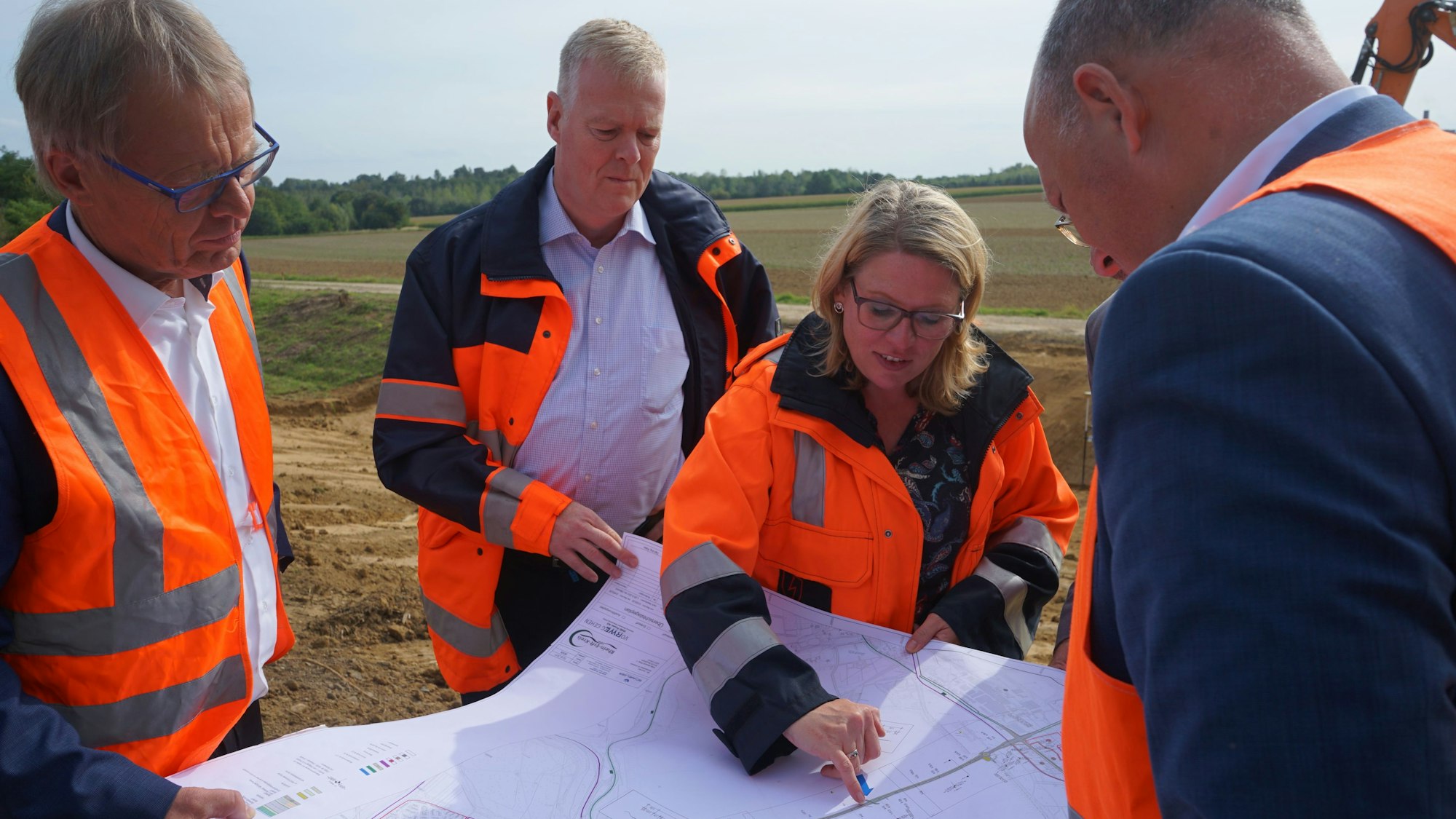 Drei Männer und eine Frau in Warnwesten beugen sich vor einem Acker über eine Karte, im Hintergrund ist der Arm eines Baggers zu erkennen.