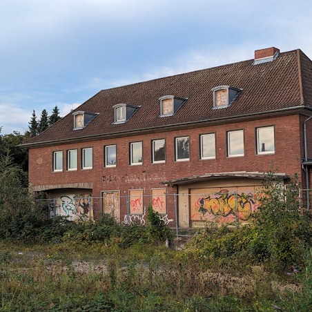 Ein Bahnhofsgebäude mit zugenagelten Fenstern und Türen hinter einem überwucherten ehemaligen Gleisbett.