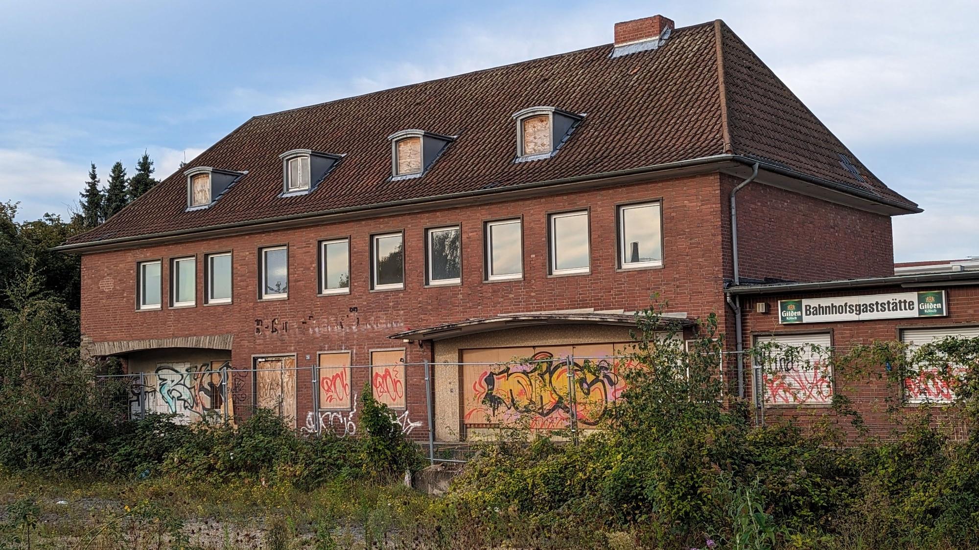 Ein Bahnhofsgebäude mit zugenagelten Fenstern und Türen hinter einem überwucherten ehemaligen Gleisbett.