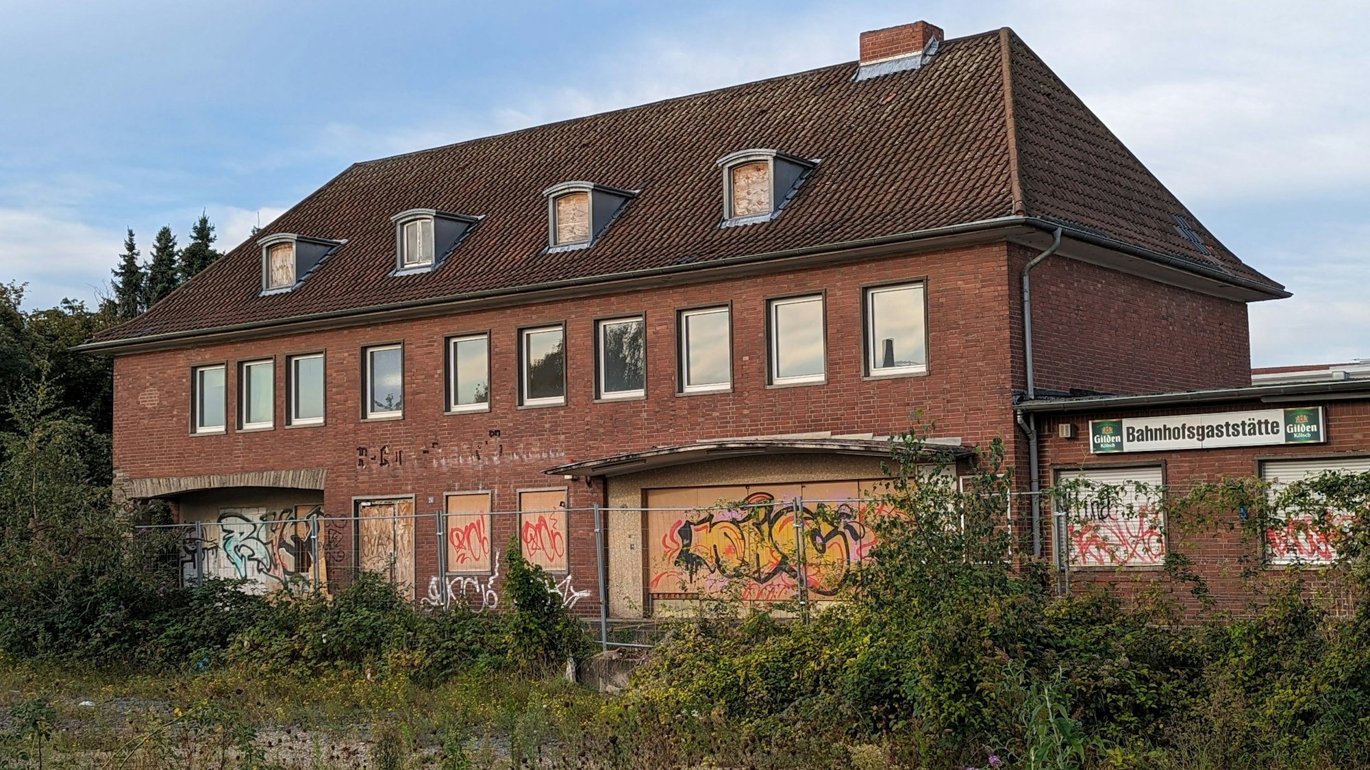 Ein Bahnhofsgebäude mit zugenagelten Fenstern und Türen hinter einem überwucherten ehemaligen Gleisbett.