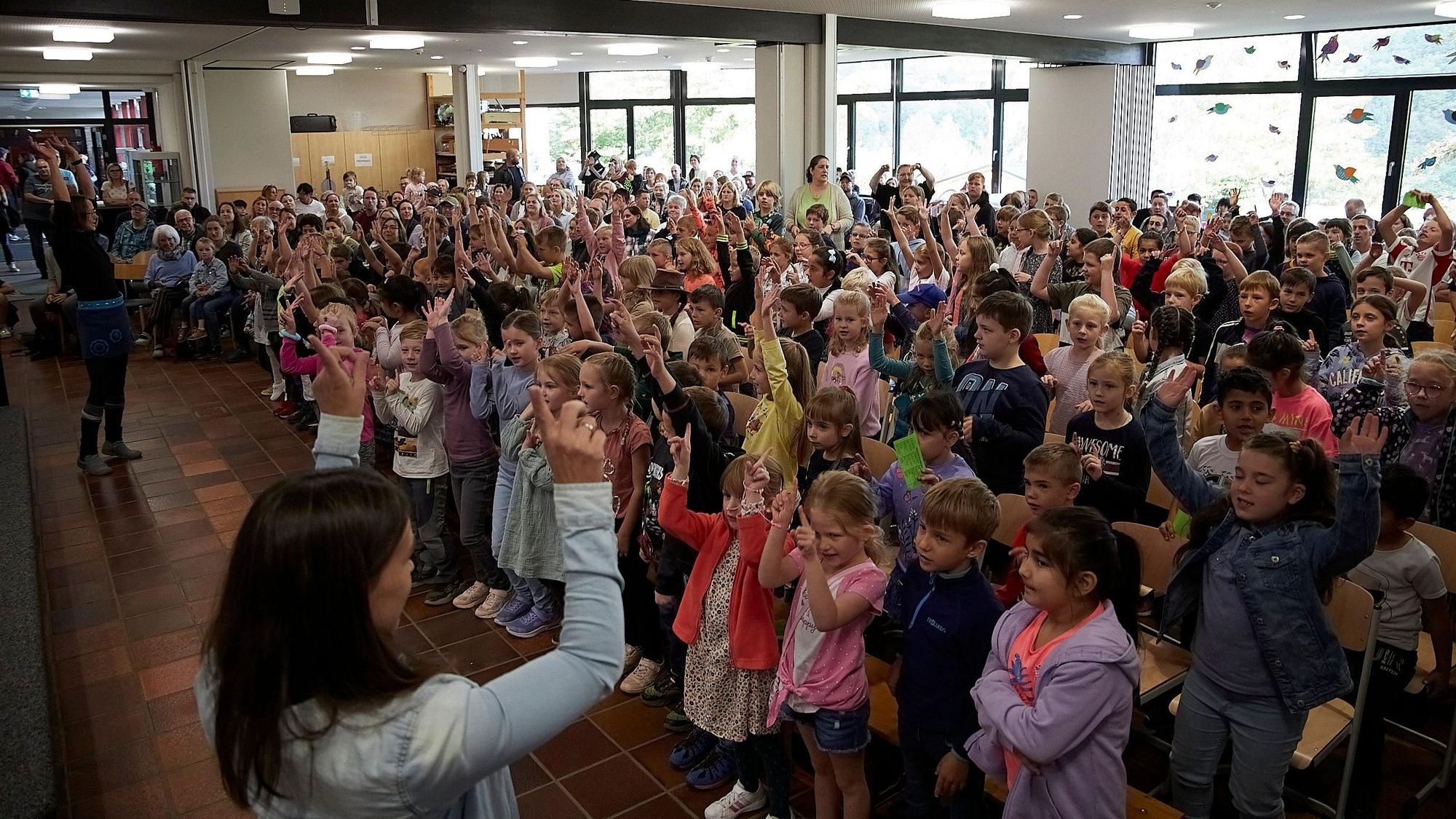 Viele Kinder stehen in der Aula der Grunschule Heimbach. Sie singen, die meisten haben eine Hand erhoben.