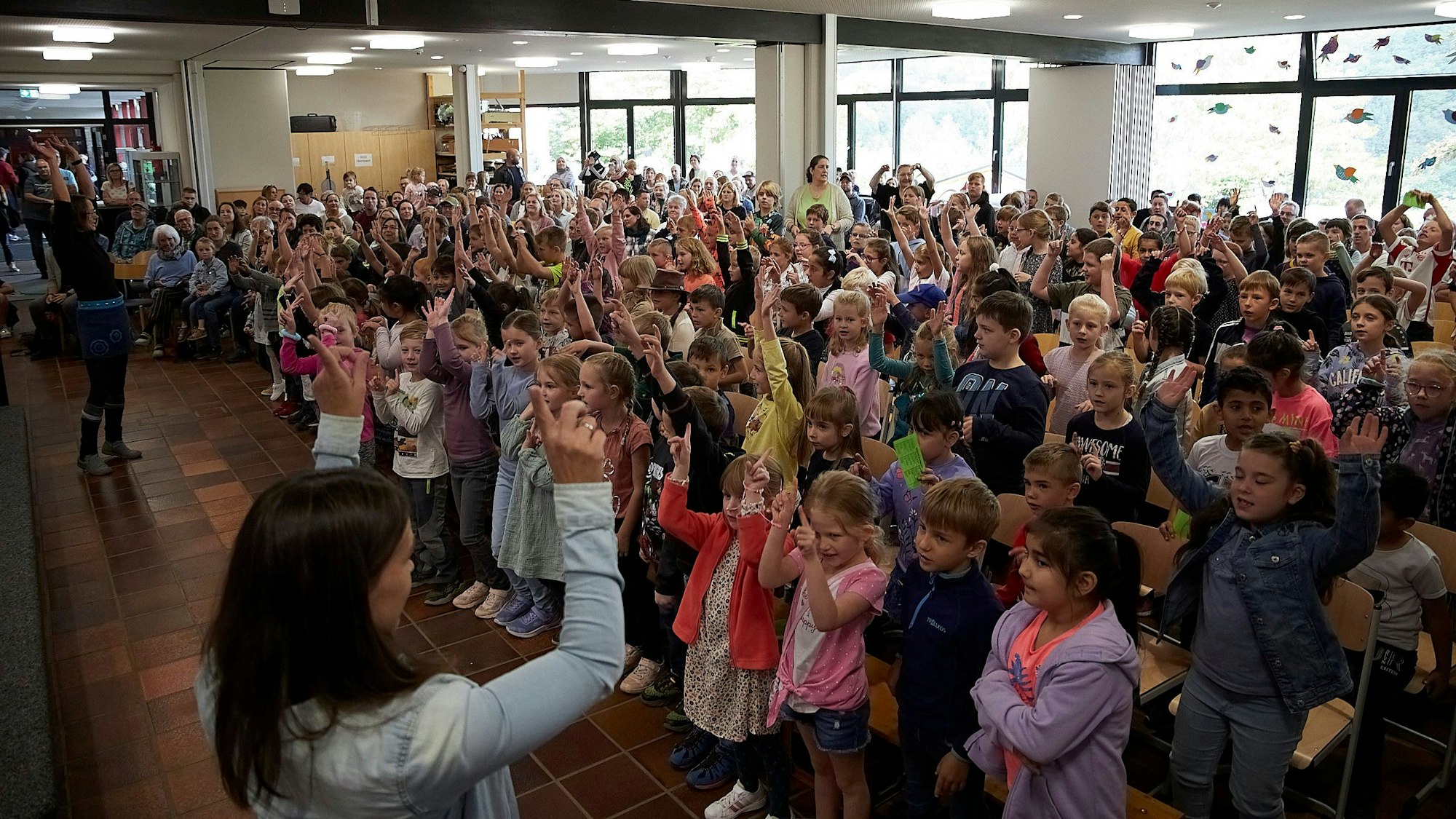 Viele Kinder stehen in der Aula der Grunschule Heimbach. Sie singen, die meisten haben eine Hand erhoben.