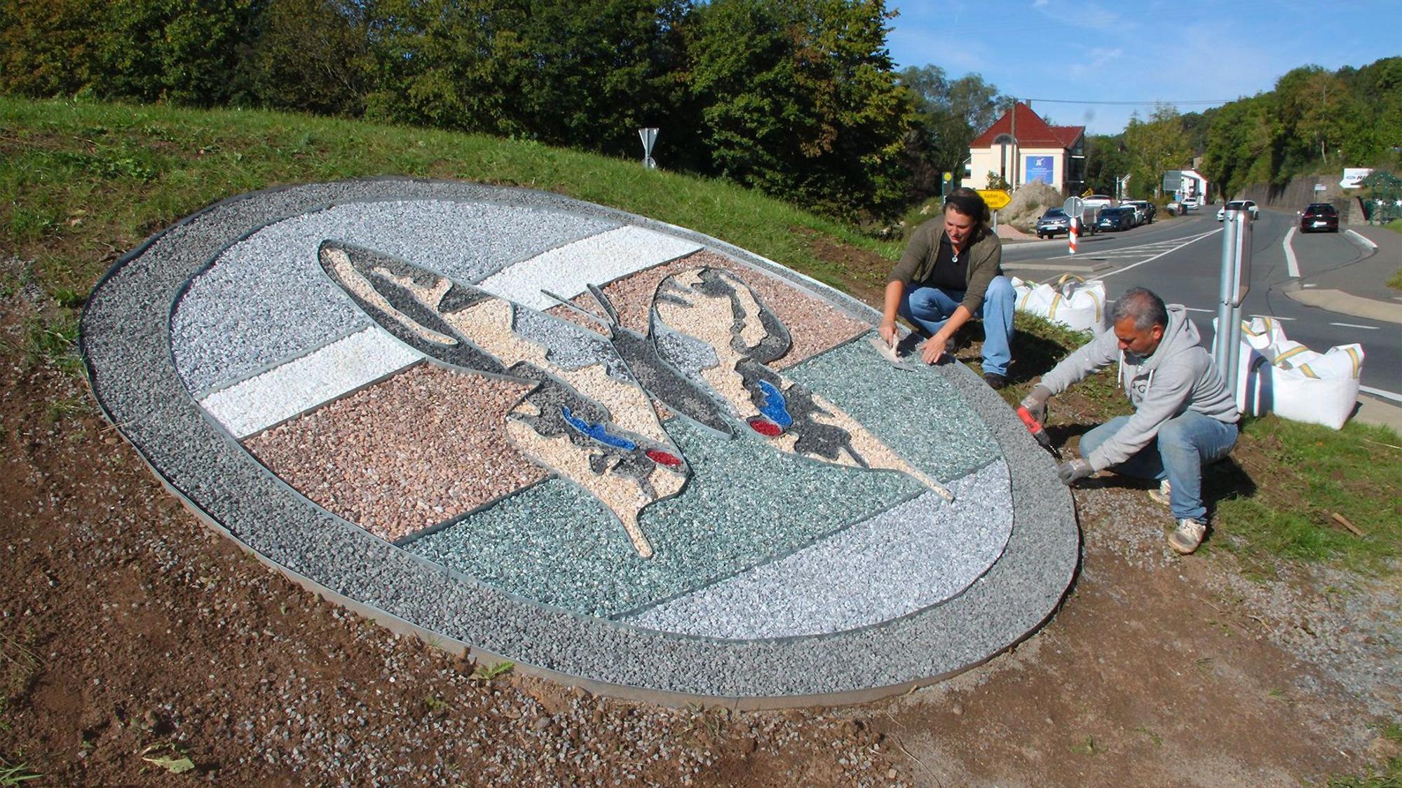 Auf der Insel des Kreisverkehrs in Ahrhütte legen ein Mann und eine Frau ein Schmetterlings-Bild aus farbigem Splitt.