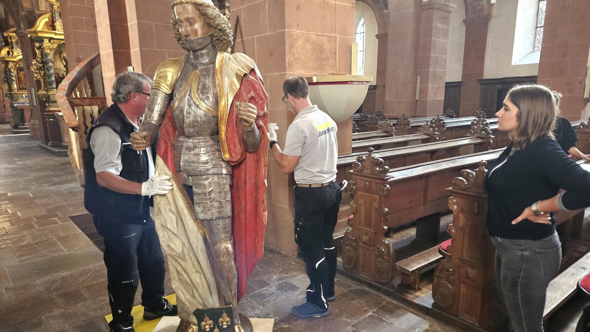 Zwei Männer transportieren die Figur des heiligen Potentinus auf einem Brett mit Rollen. Die Restauratorin Sarah Renn schaut zu.