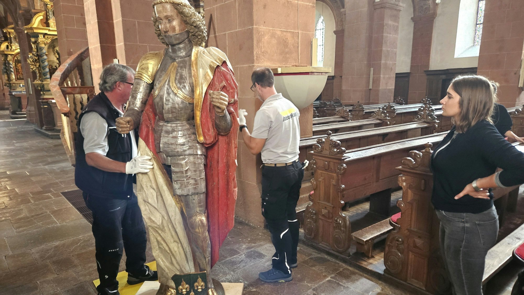 Zwei Männer transportieren die Figur des heiligen Potentinus auf einem Brett mit Rollen. Die Restauratorin Sarah Renn schaut zu.