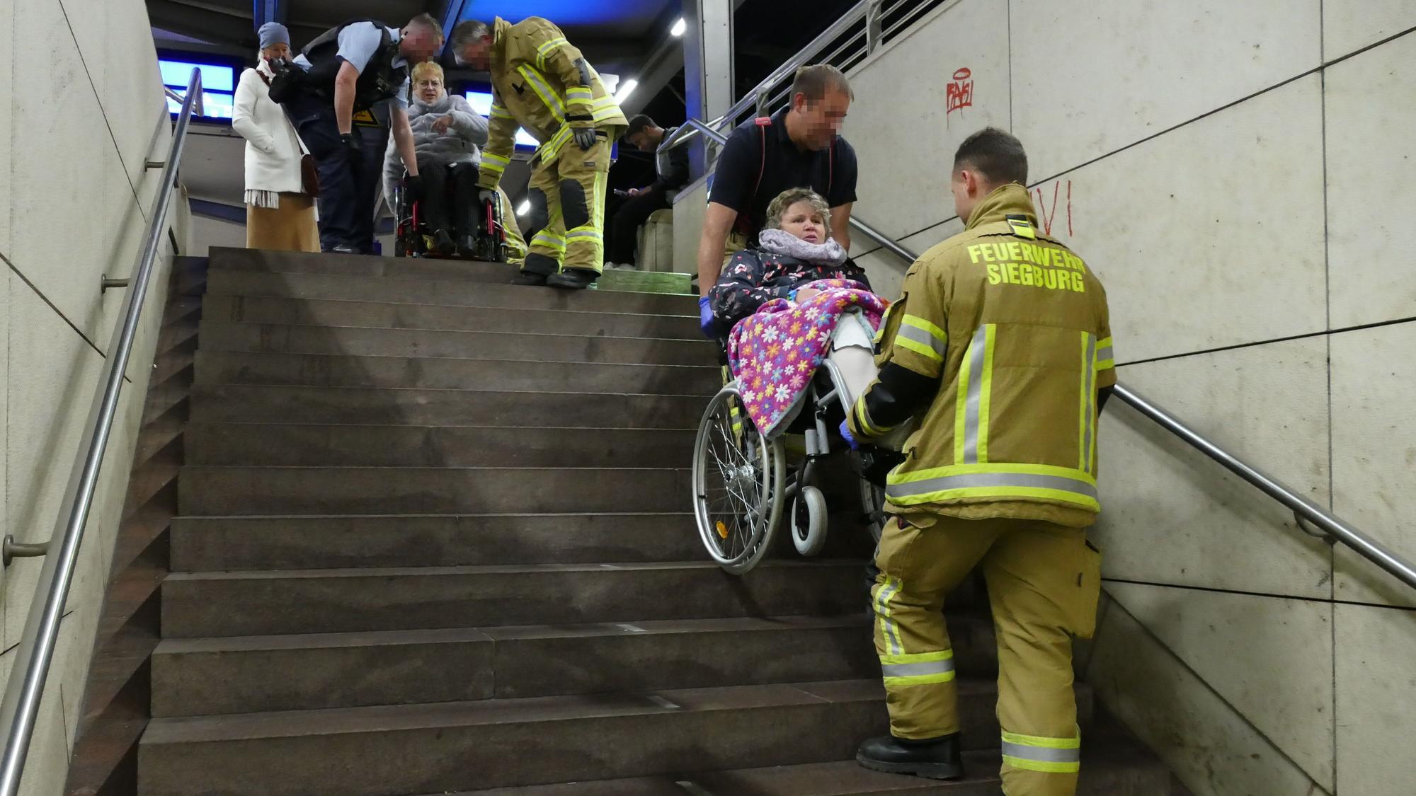 Zwei Männer in gelben Feuerwehranzügen tragen zwei Frauen im Rollstuhl die Treppe herunter.