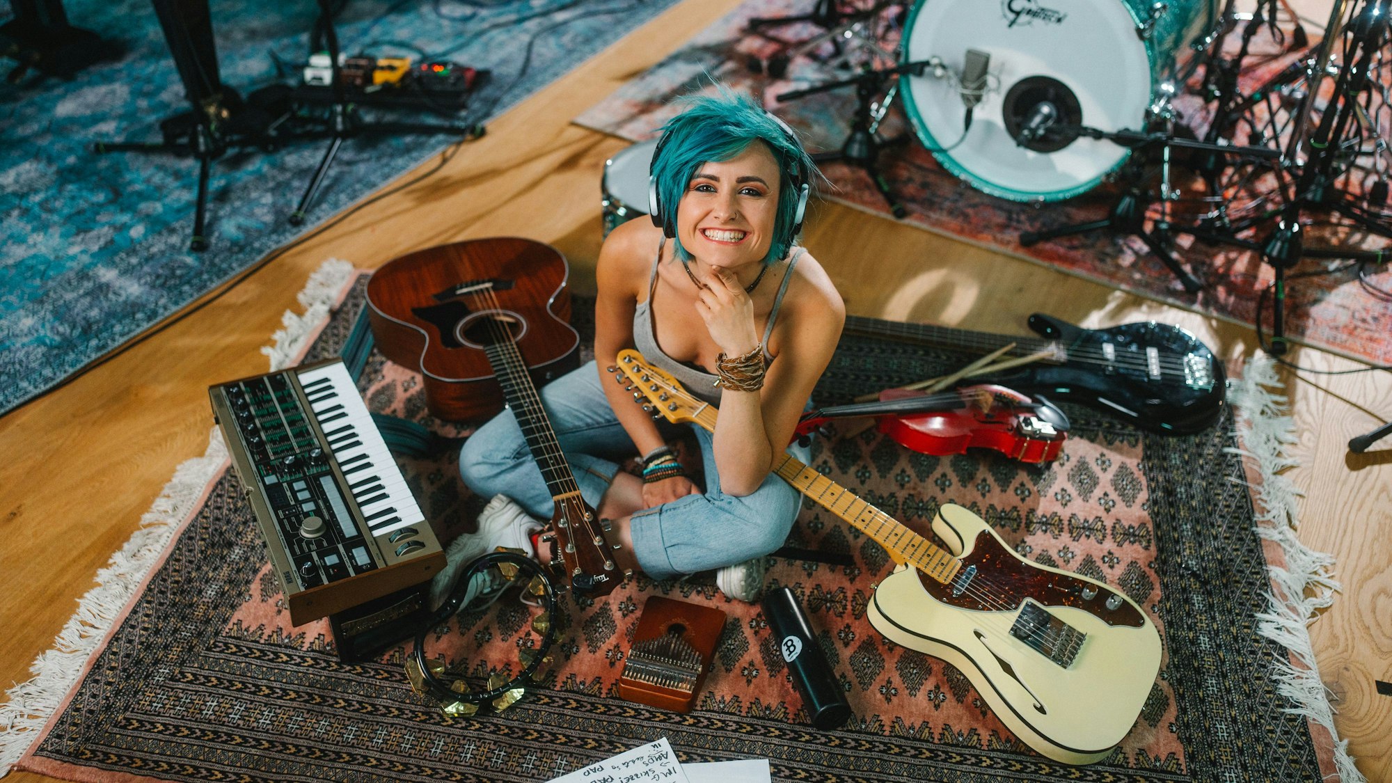 Die Musikerin Charly Klauser sitzt mit Kopfhörern auf einem Teppich, um sie herum sind Rockmusikinstrumente drapiert.