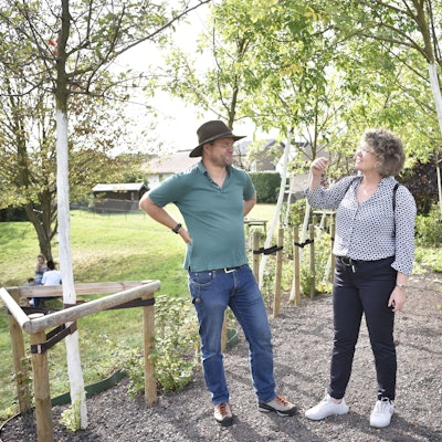 Ein Mann und eine Frau, Christoph Zimmermann und Anne Horst, stehen unter einer Reihe junger Bäume und unterhalten sich.