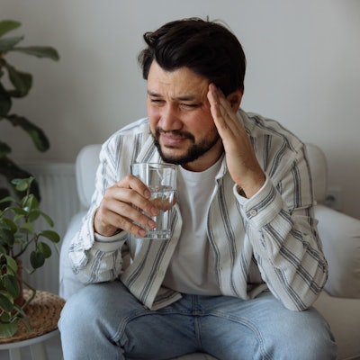 Ein Mann sitzt mit verzerrtem Gesicht im Wohnzimmer, hält mit der einen Hand seinen Kopf und in der anderen hand ein Glas.