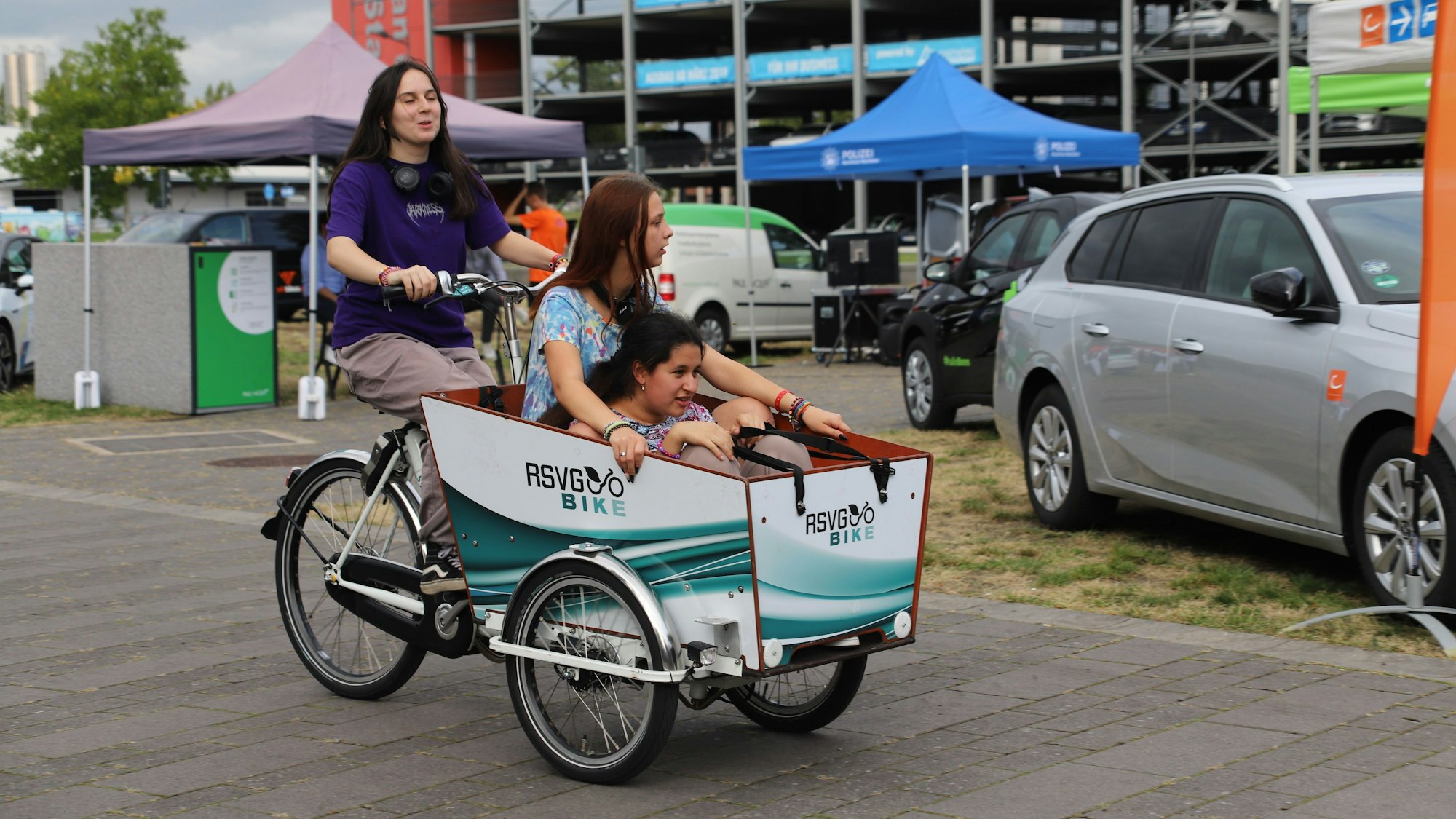 Auf einem Fahrrad sitzt eine junge Frau, im Lastenkorb davor sitzen zwei weitere junge Frauen..
