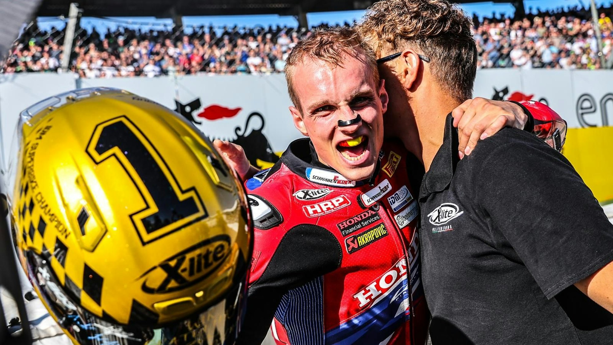 Florian Alt jubelt und umarmt einen Teamkollegen. In der Hand hält er einen goldenen Motorradhelm.