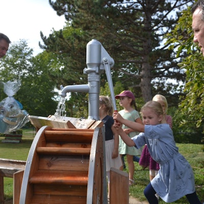 Euskirchens Bürgermeister Sacha Reichelt (r.) und Stiftungsgeschäftsführer Christoph Palm (l.) schauen zu, wie die Kinder der Kita Großbüllesheim die Wassermatschanlage bedienen. Aus einer Pumpe läuft Wasser.