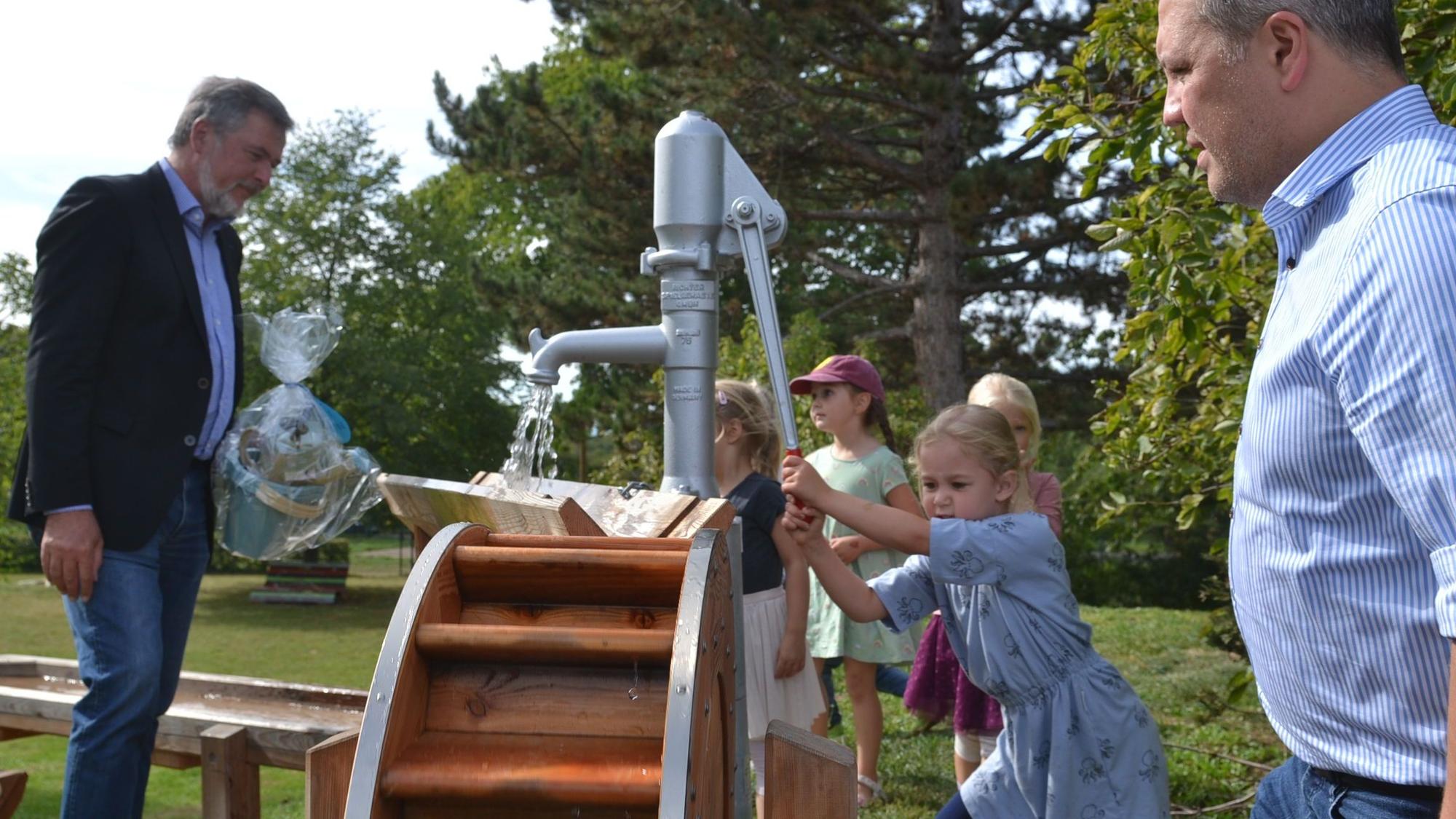 Euskirchens Bürgermeister Sacha Reichelt (r.) und Stiftungsgeschäftsführer Christoph Palm (l.) schauen zu, wie die Kinder der Kita Großbüllesheim die Wassermatschanlage bedienen. Aus einer Pumpe läuft Wasser.