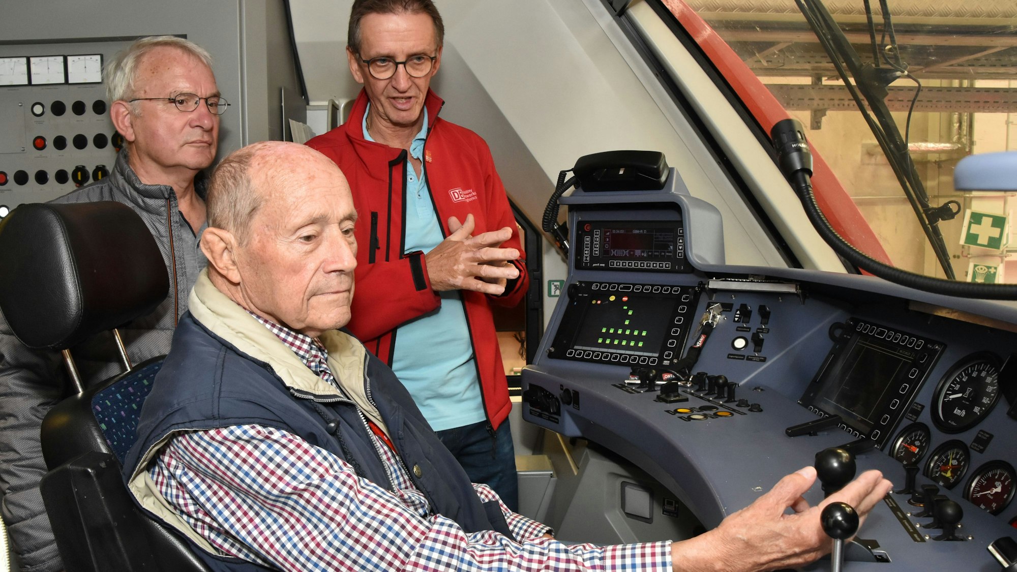 Ein Teamleiter in Roter Jacke erklärt im Führerstand einer Lok einem Senior die Technik.