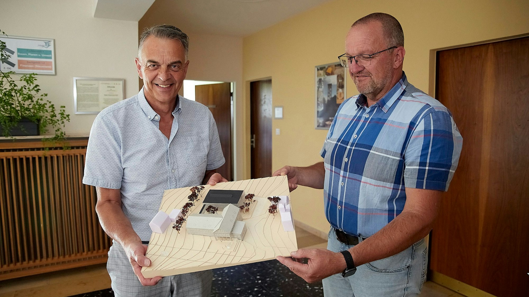 Bürgermeister Rudolf Westerburg (links) und Bauamtsleiter Markus Rodenbüsch stehen in einem Flur des Hellenthaler Rathauses. Sie halten ein Modell in den Händen, das vorne das bestehende Rathaus zeigt und dahinter, mit schwarzem Dach, den geplanten Anbau.
