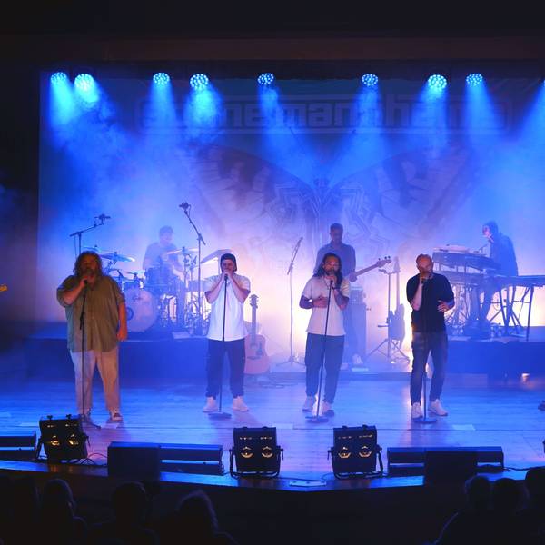 Eine Band mit vier Sängern im Vordergrund und mehreren Instrumentalisten steht auf der blau illuminierten Bühne des Euskirchener Theaters.