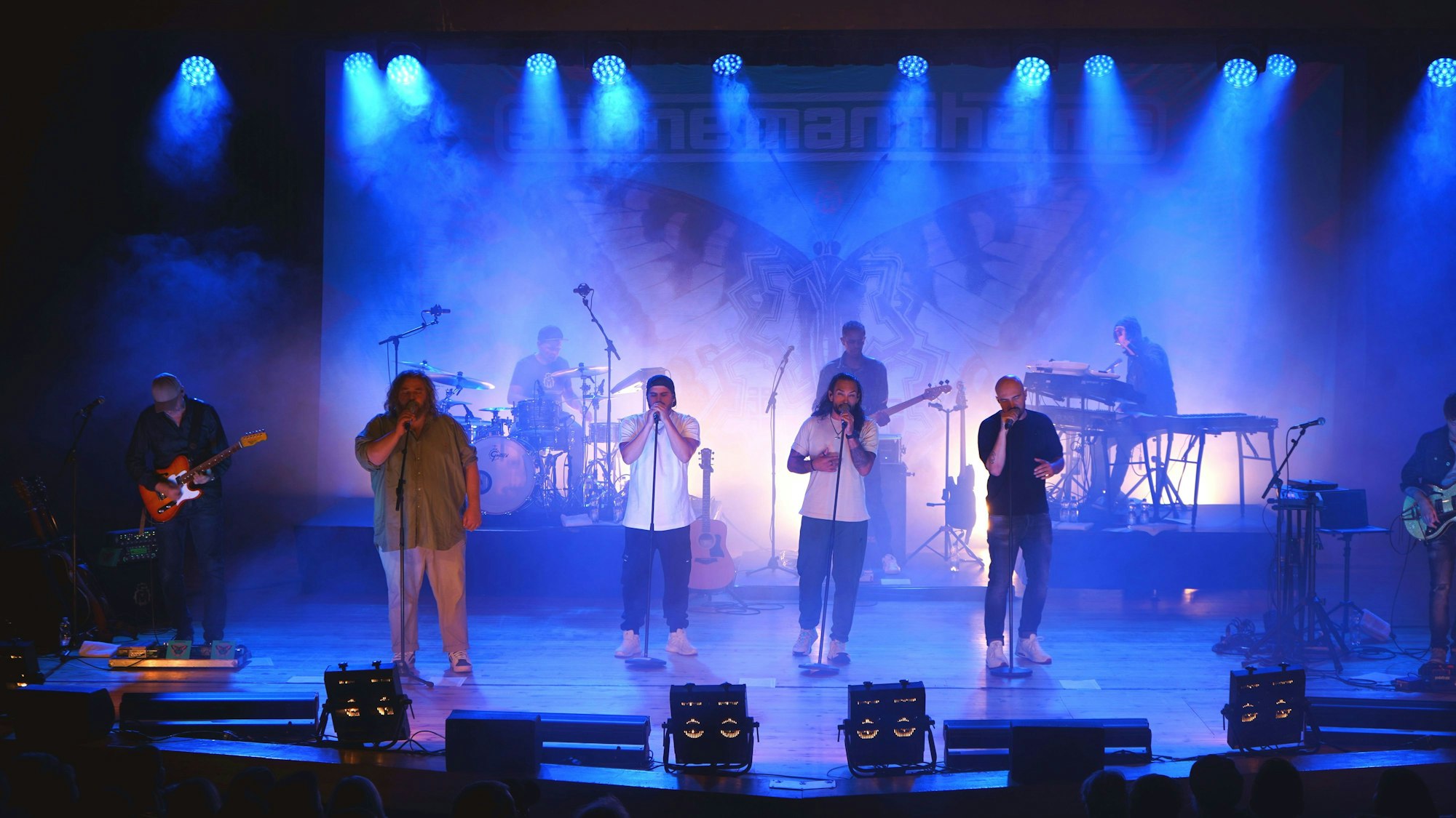 Eine Band mit vier Sängern im Vordergrund und mehreren Instrumentalisten steht auf der blau illuminierten Bühne des Euskirchener Theaters.