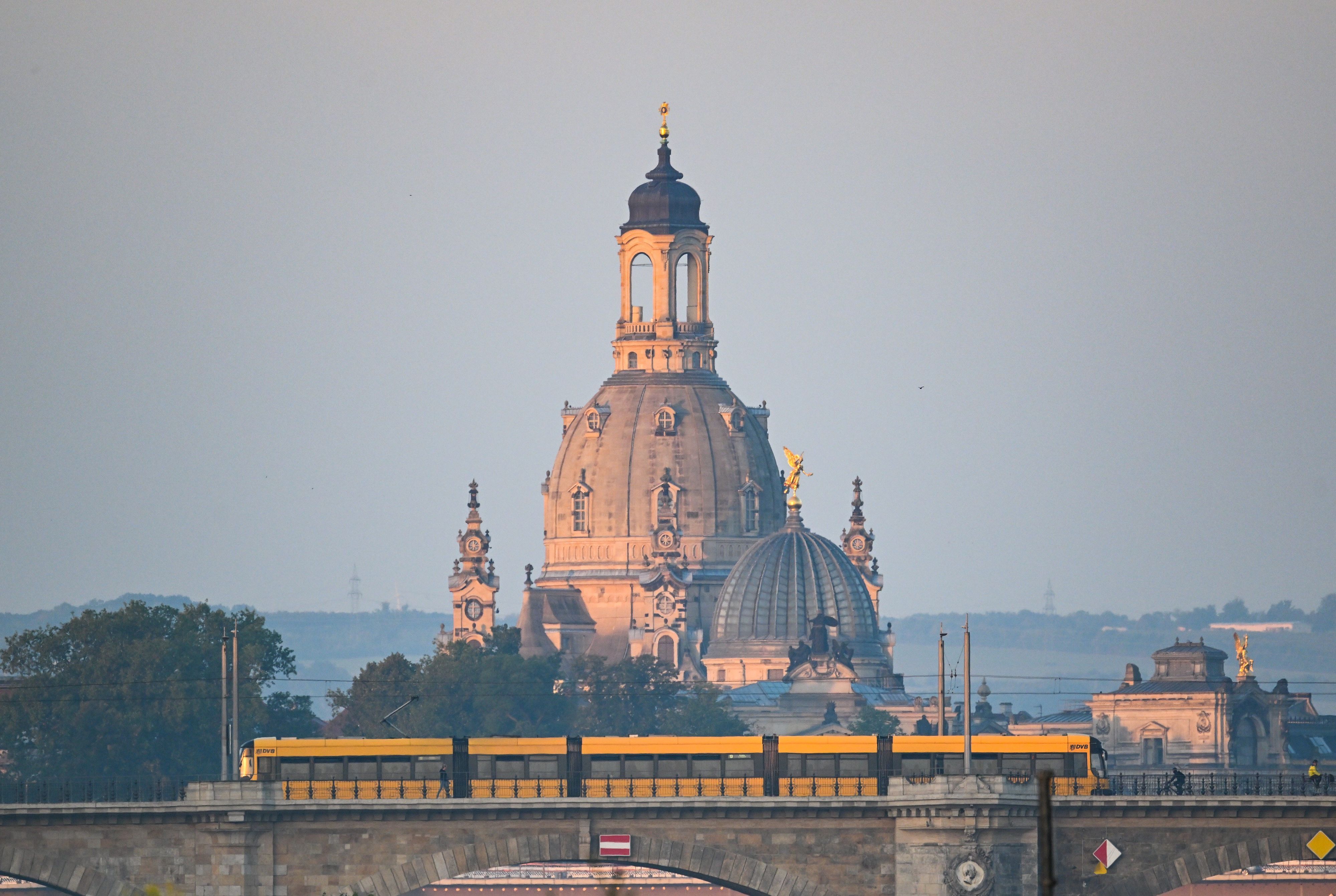 Eine Straßenbahn der Dresdner Verkehrsbetriebe (DVB) fährt am Morgen vor der Altstadtkulisse mit der Frauenkirche (l) und der Kuppel der Kunstakedmie mit dem Engel «Fama» über die Albertbrücke.