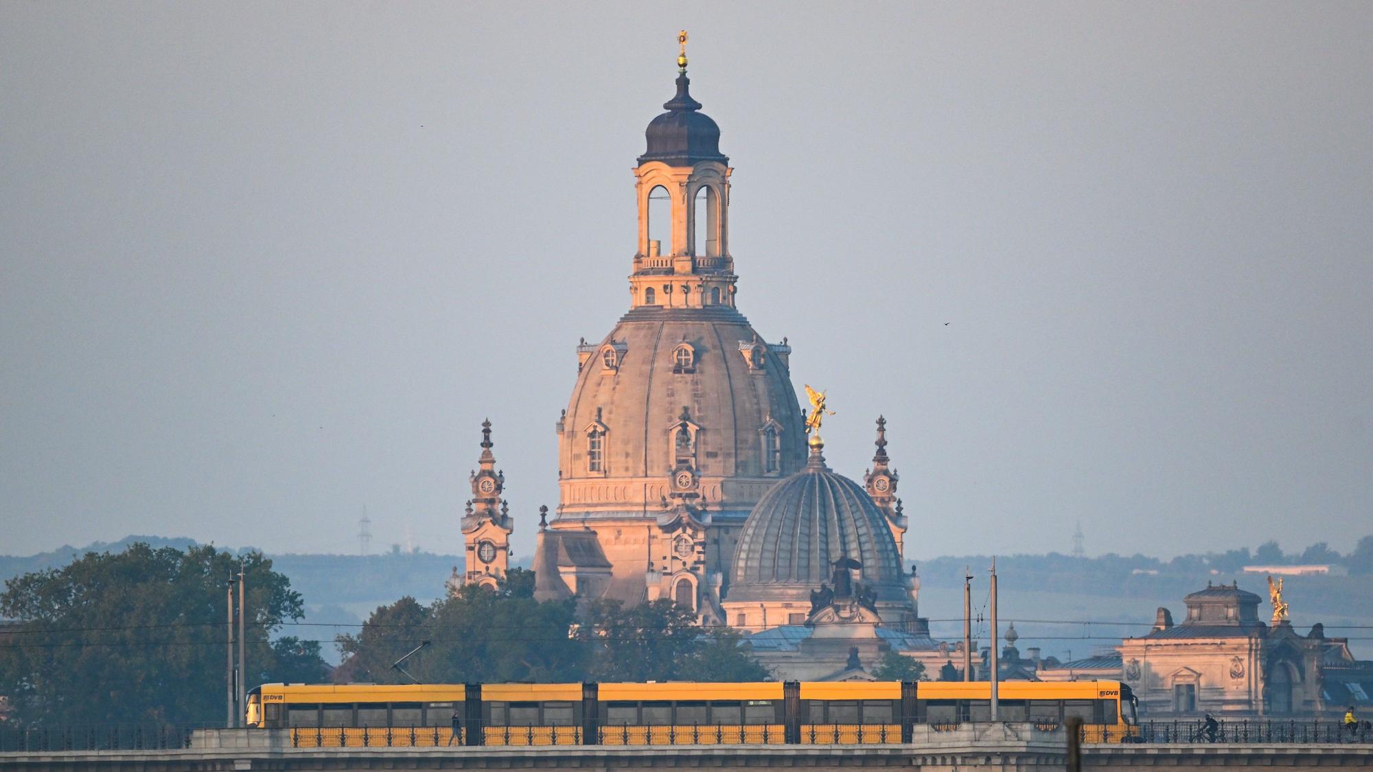 Eine Straßenbahn der Dresdner Verkehrsbetriebe (DVB) fährt am Morgen vor der Altstadtkulisse mit der Frauenkirche (l) und der Kuppel der Kunstakedmie mit dem Engel «Fama» über die Albertbrücke.