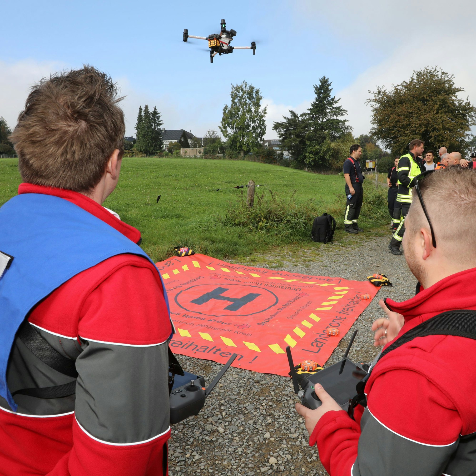 Zwei DRK-Einsatzkräfte stehen mit Fernbedienungen an einem roten Tuch mit einem großen „H“, von dem gerade eine Drohne abgehoben hat.