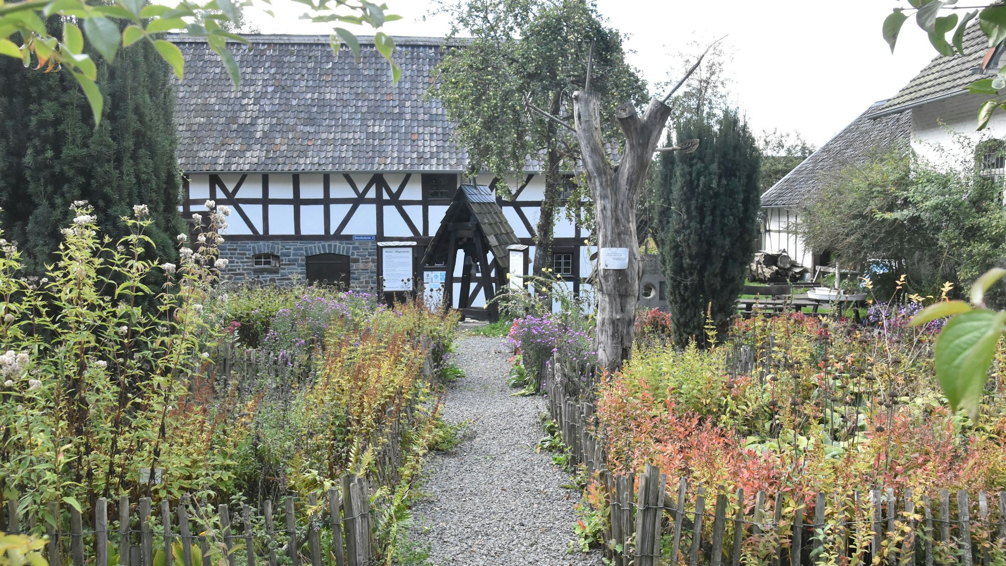 Fachwerkhäuser im Hintergrund, davor ein mit einem Staketenzaun eingefasster Bauerngarten, der von einem Kiesweg durchschnitten wird.