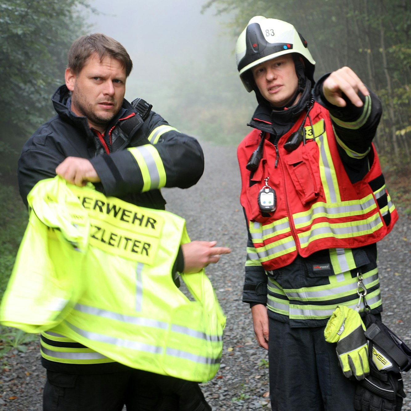 Ein Feuerwehrmann zieht eine gelbe Weste mit der Aufschrift „Feuerwehr Einsatzleiter“ an, ein anderer erklärt ihm etwas und deutet nach vorne.