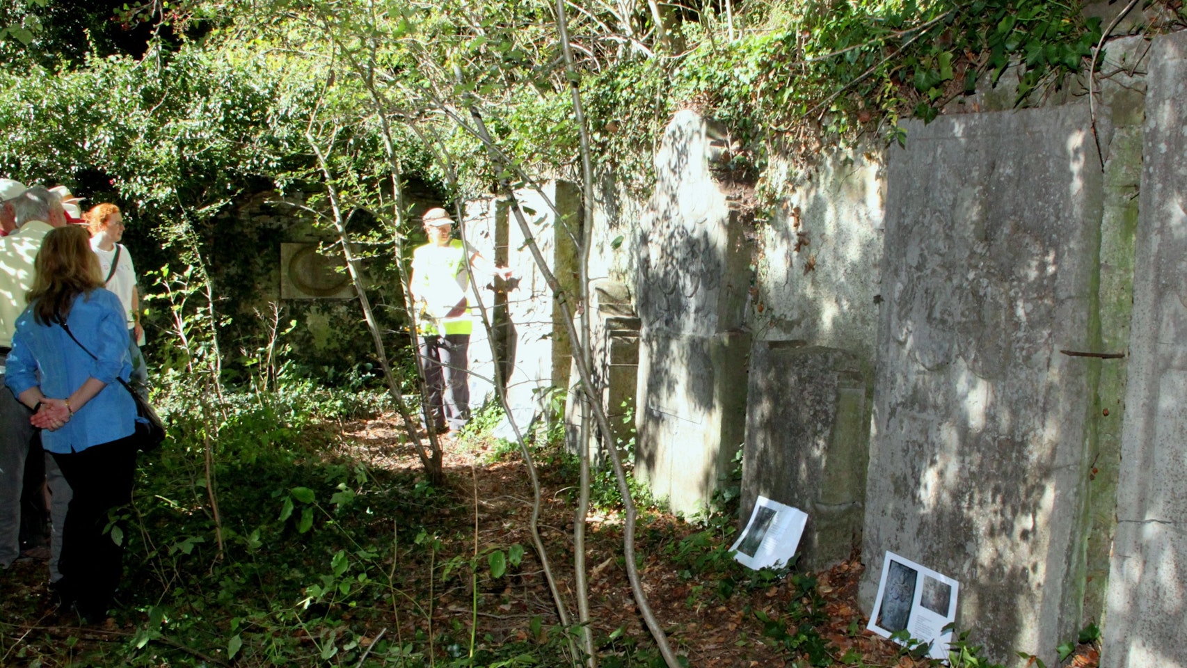Auf der rechten Seite des Bildes sind Steinplatten eines alten Friedhofs zu sehen, die mit grünen Pflanzen teils überwachsen sind. Links stehen einige Menschen, die das Ensemble an der Burg Dalbenden betrachten.