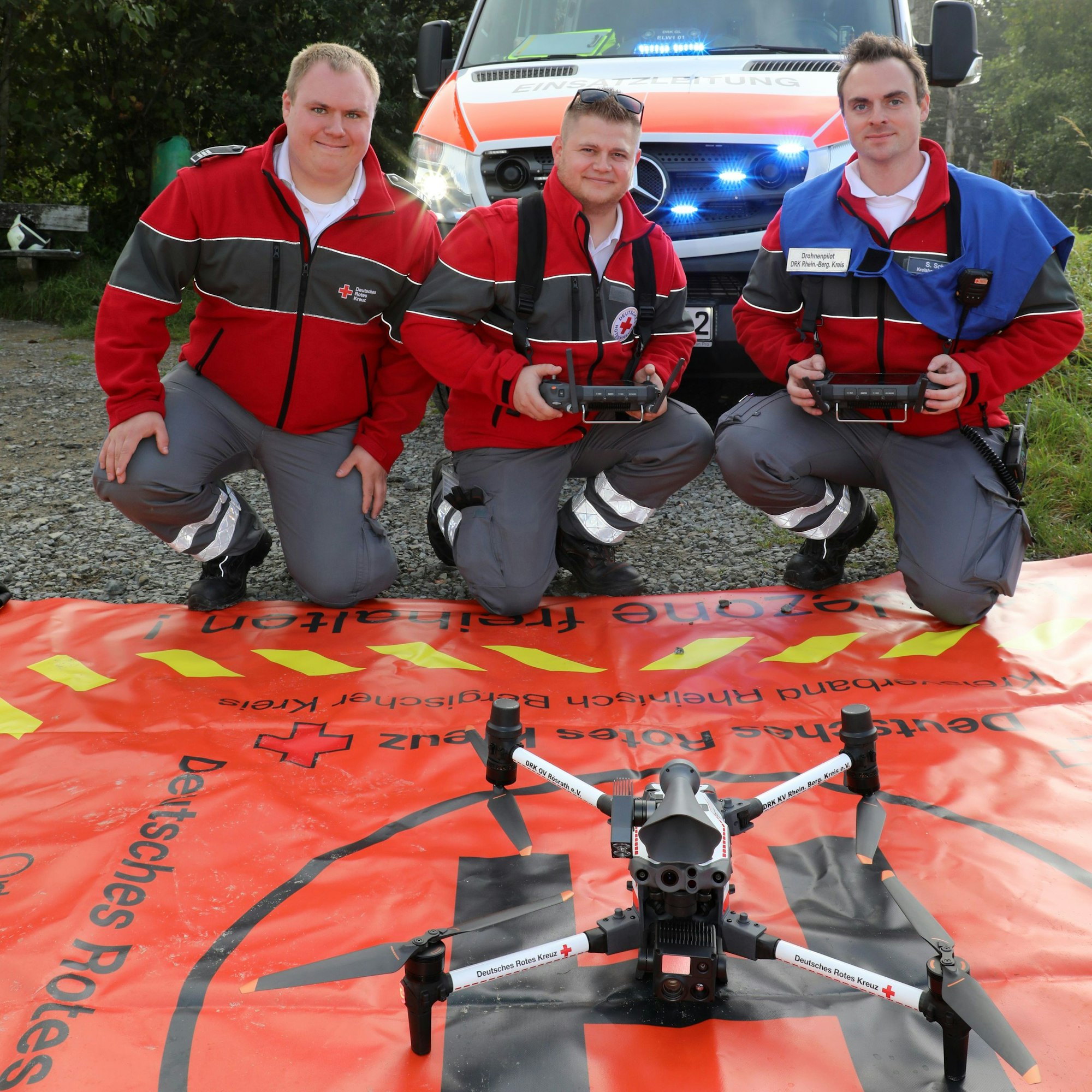 Drei DRK-Helfer mit Fernbedienungen hocken vor einer großen Drohne, die auf einem Start- und Landesplatz-Tuch steht.