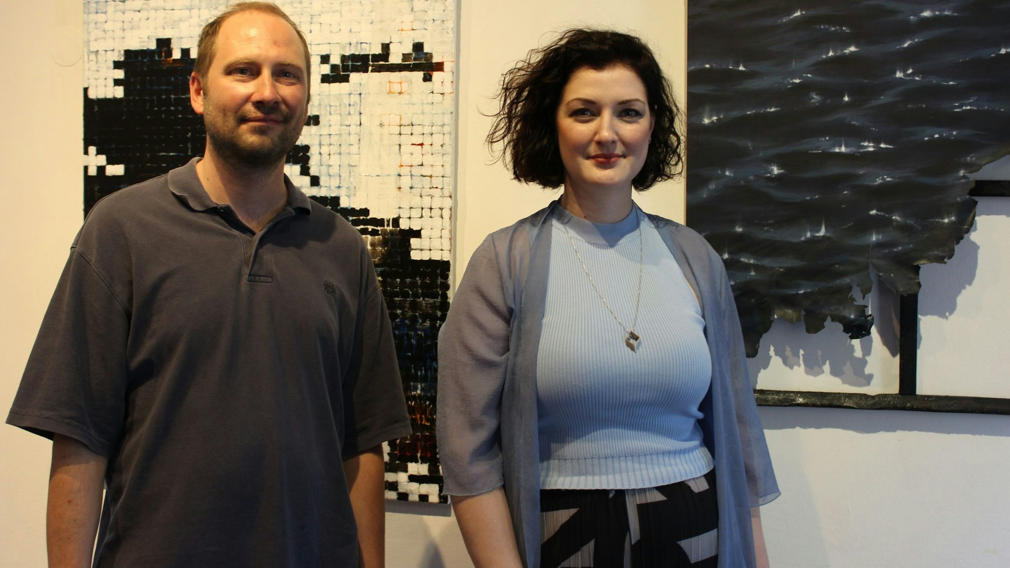 Die Künstlerin und der Künstlerin stehen vor zwei abstrakt wirkenden Gemälden.