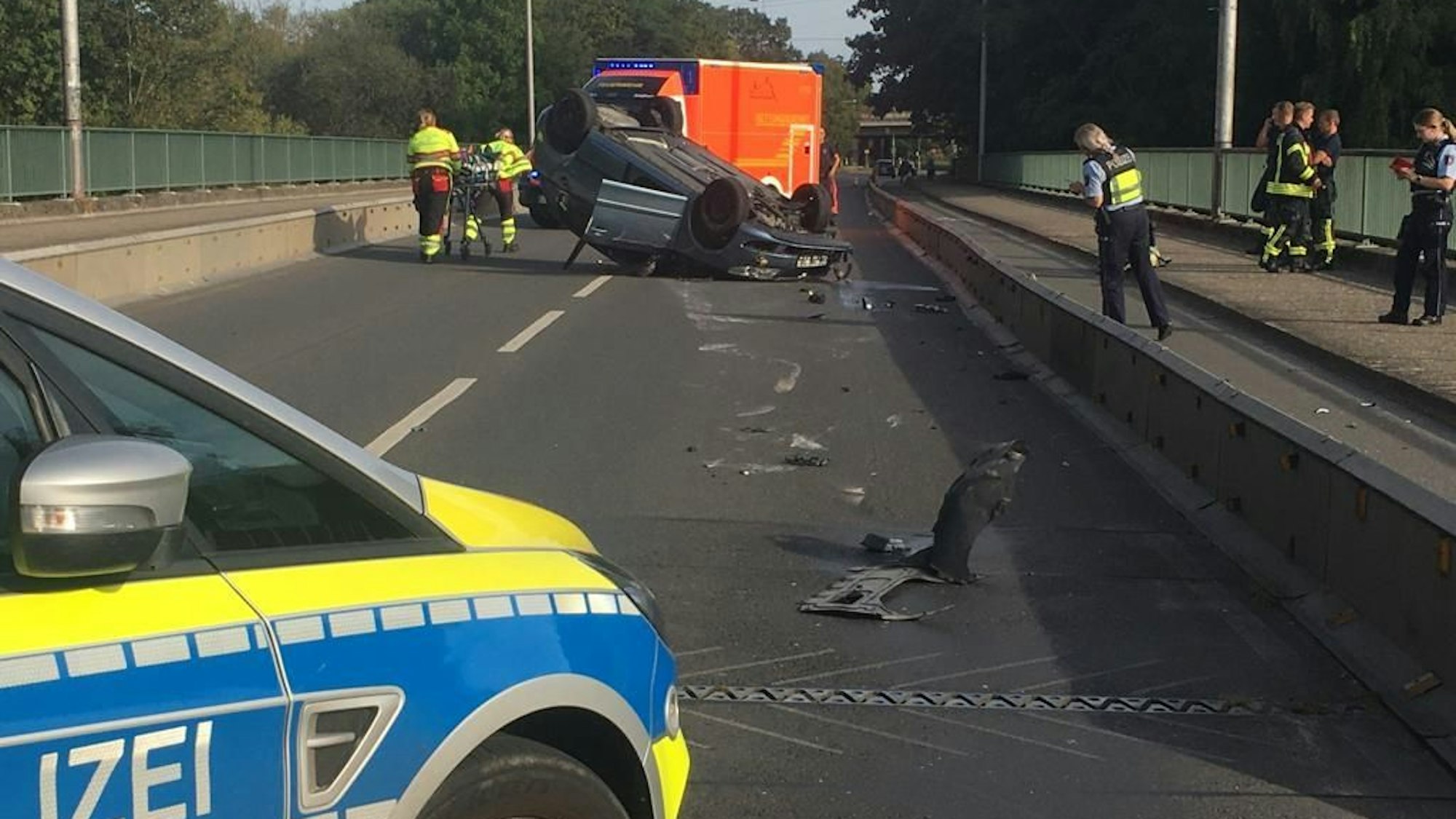 Bei einem Unfall in Troisdorf, bei dem sich ein Auto überschlug, wurde ein 56 Jahre alter Mann verletzt. Das Auto liegt auf dem Dach, Mitarbeiterinnen des Rettungsdienstes bringen den Autofahrer zu einem Rettungswagen. Polizistinnen sichern Spuren.