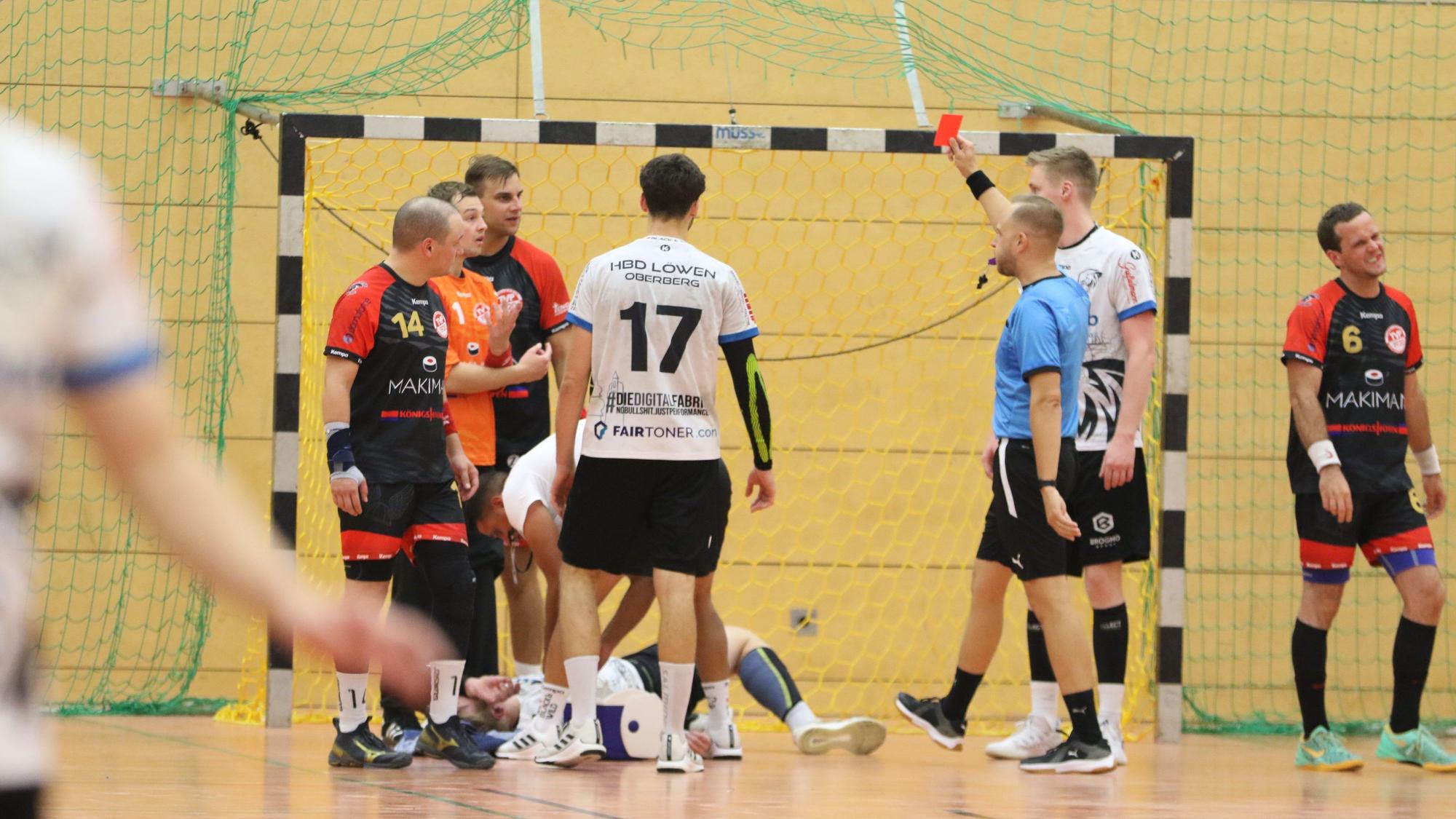 Ein Schiedsrichter zeigt einem Handballspieler die Rote Karte. Die Mitspieler sind fassungslos. Ein gegnerischer Spieler wird behandelt.