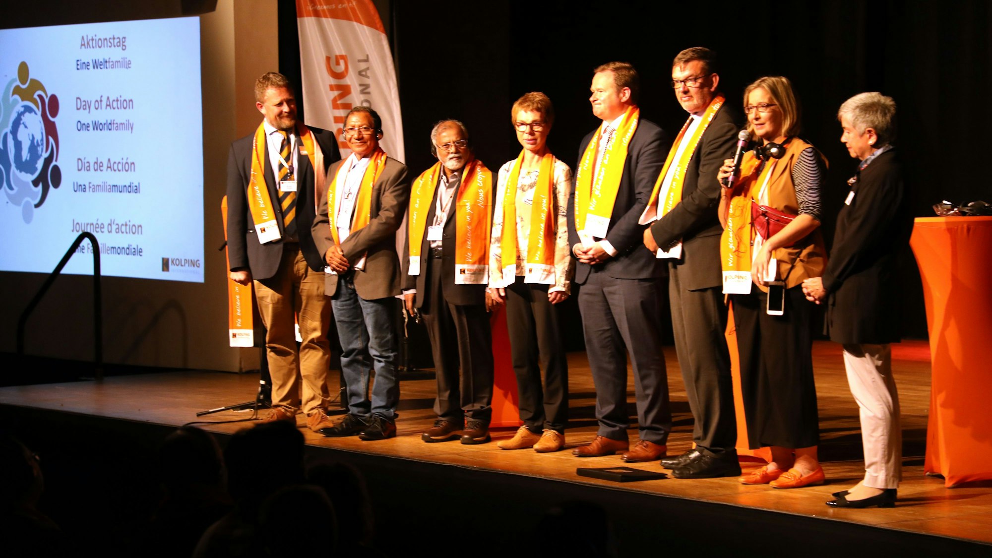 Menschen stehen auf einer Bühne, eine Moderatorin hat ein Mikrofon in der Hand.