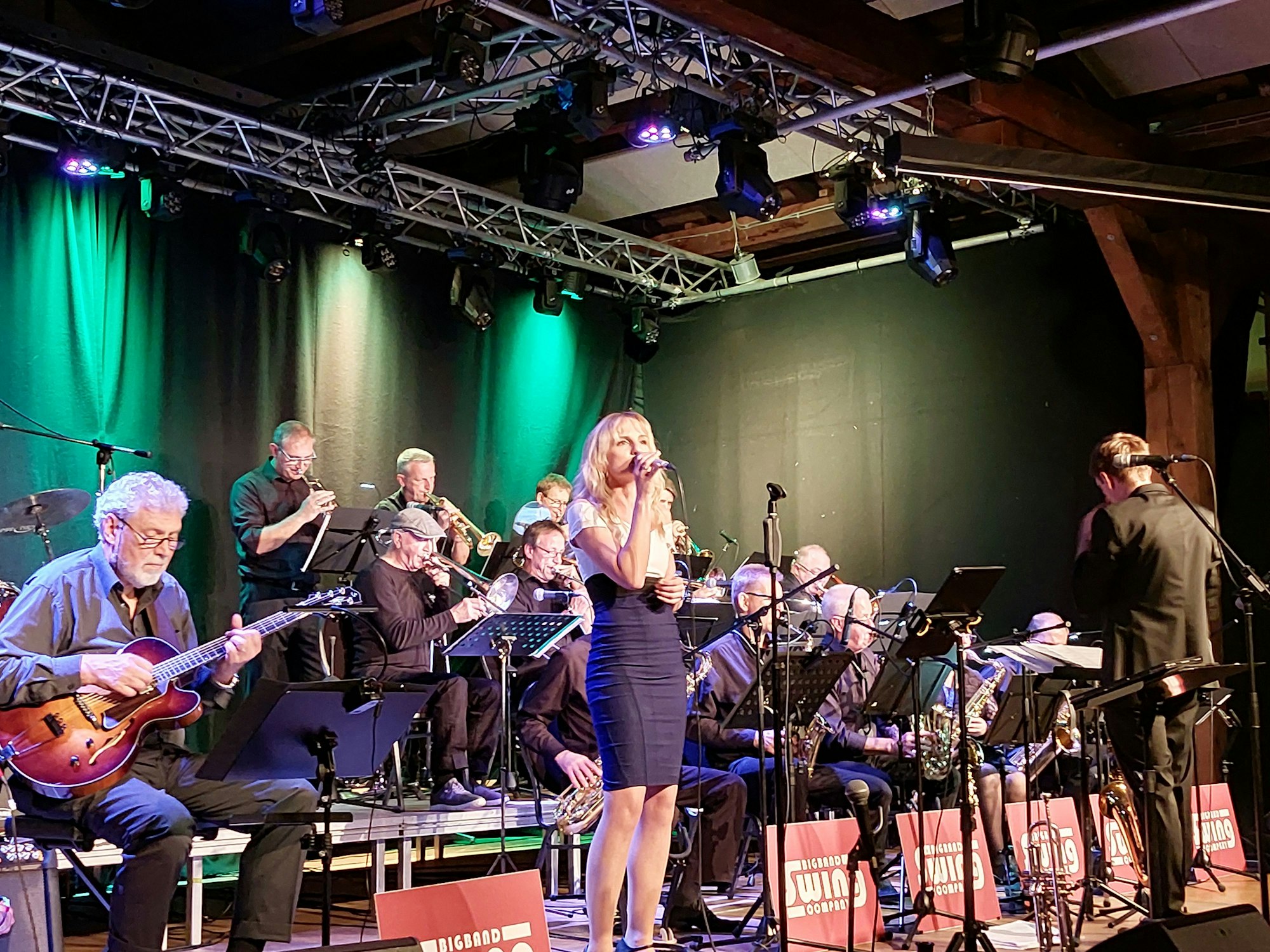 Eine blonde Frau in einem engen, schwarz-weiße Kleid steht an einem Mikrofon auf der Bühne, hinter ihr sitzt die BigBand.