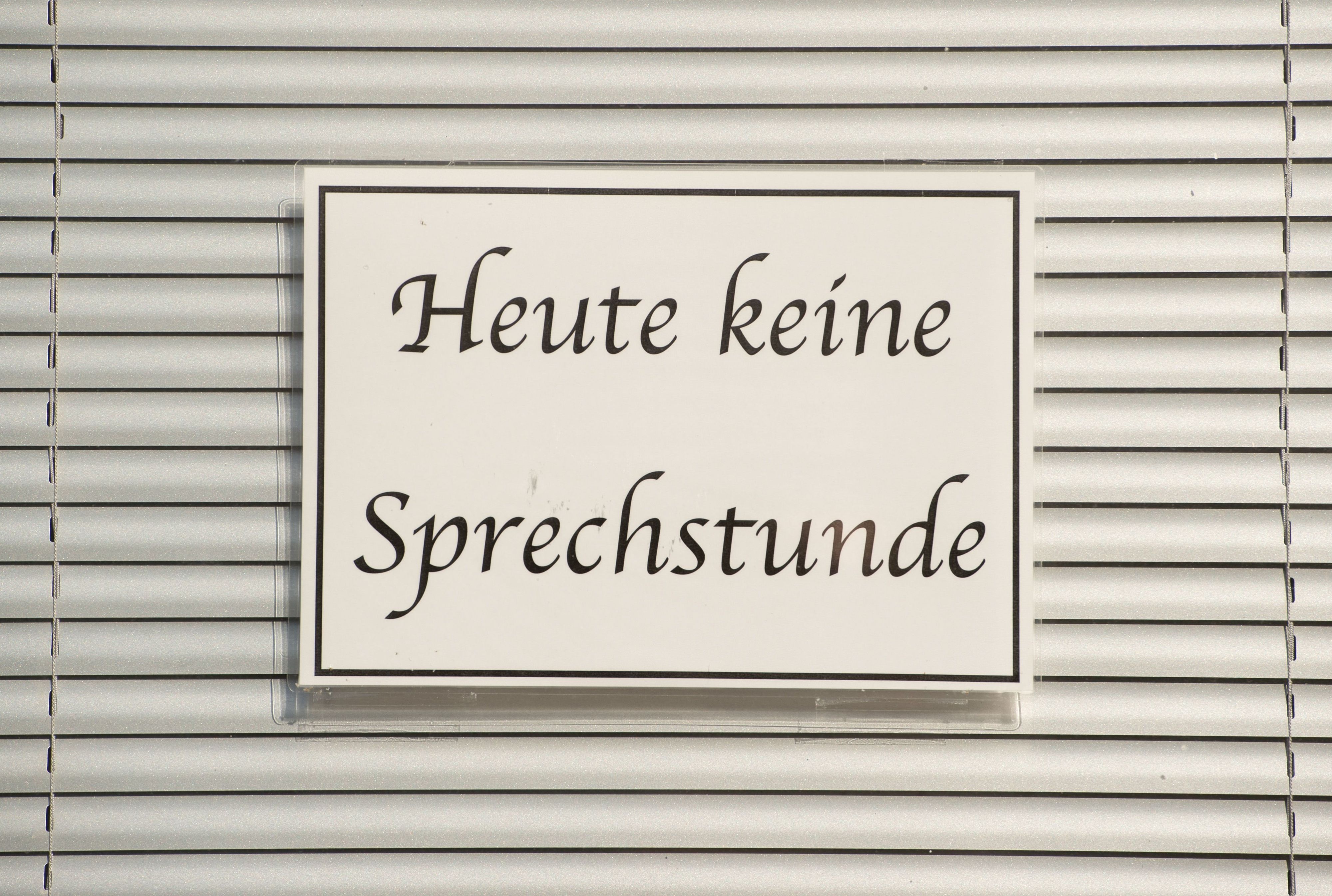 Ein Schild mit der Aufschrift ‚Heute keine Sprechstunde‘ hängt im Fenster einer Arztpraxis vor einer Jalousie.