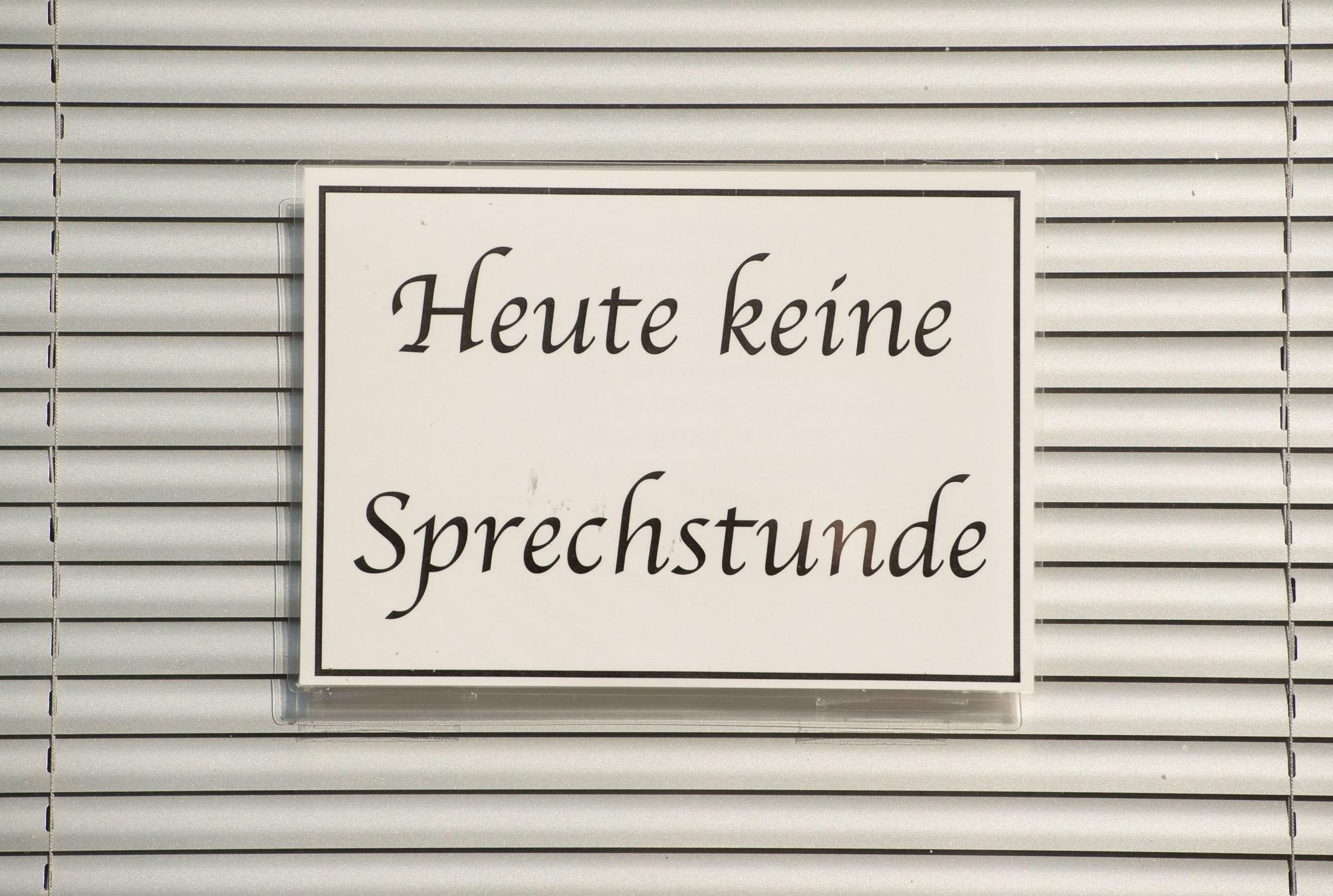 Ein Schild mit der Aufschrift ‚Heute keine Sprechstunde‘ hängt im Fenster einer Arztpraxis vor einer Jalousie.