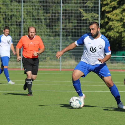 Ein Fußballer führt den Ball, ein Gegner hat ihn im Visier. Der Schiedsrichter beobachtet die Szene.