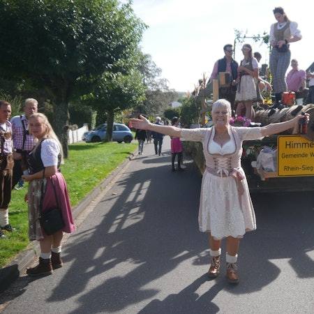 Eine Frau im Dirndl geht vor einem geschmückten Wagen, an dem das Ortsschild von Himmeroth in Windeck hängt.