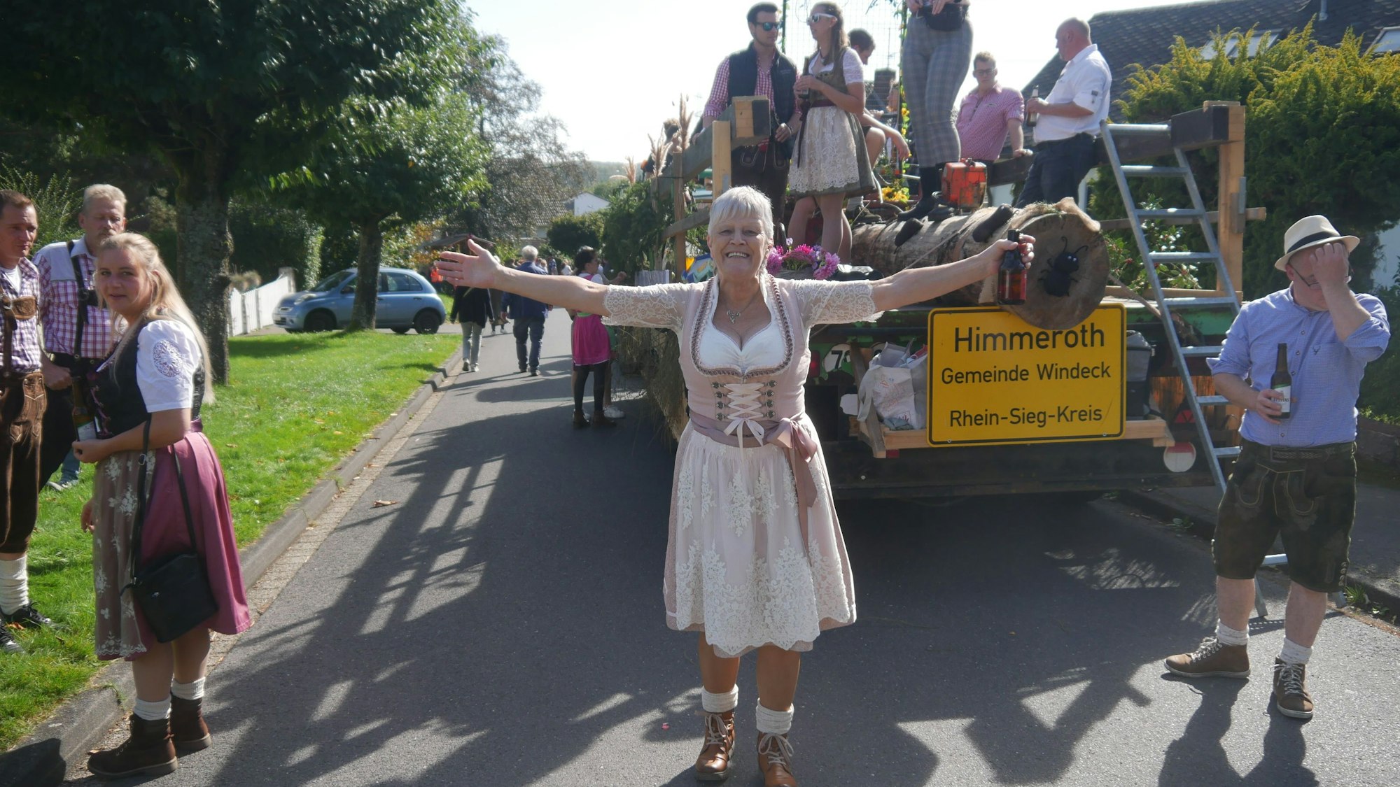 Eine Frau im Dirndl geht vor einem geschmückten Wagen, an dem das Ortsschild von Himmeroth in Windeck hängt.