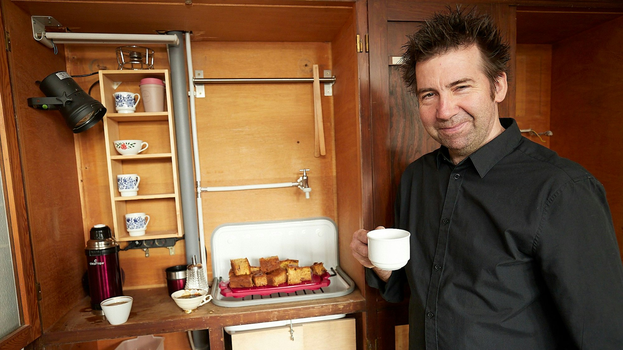 Der Künstler Rochus Aust hält eine Kaffeetasse in der Hand. Hinter ihm sind weitere Tassen und eine Platte mit Kuchen in einem Schrank zu sehen.