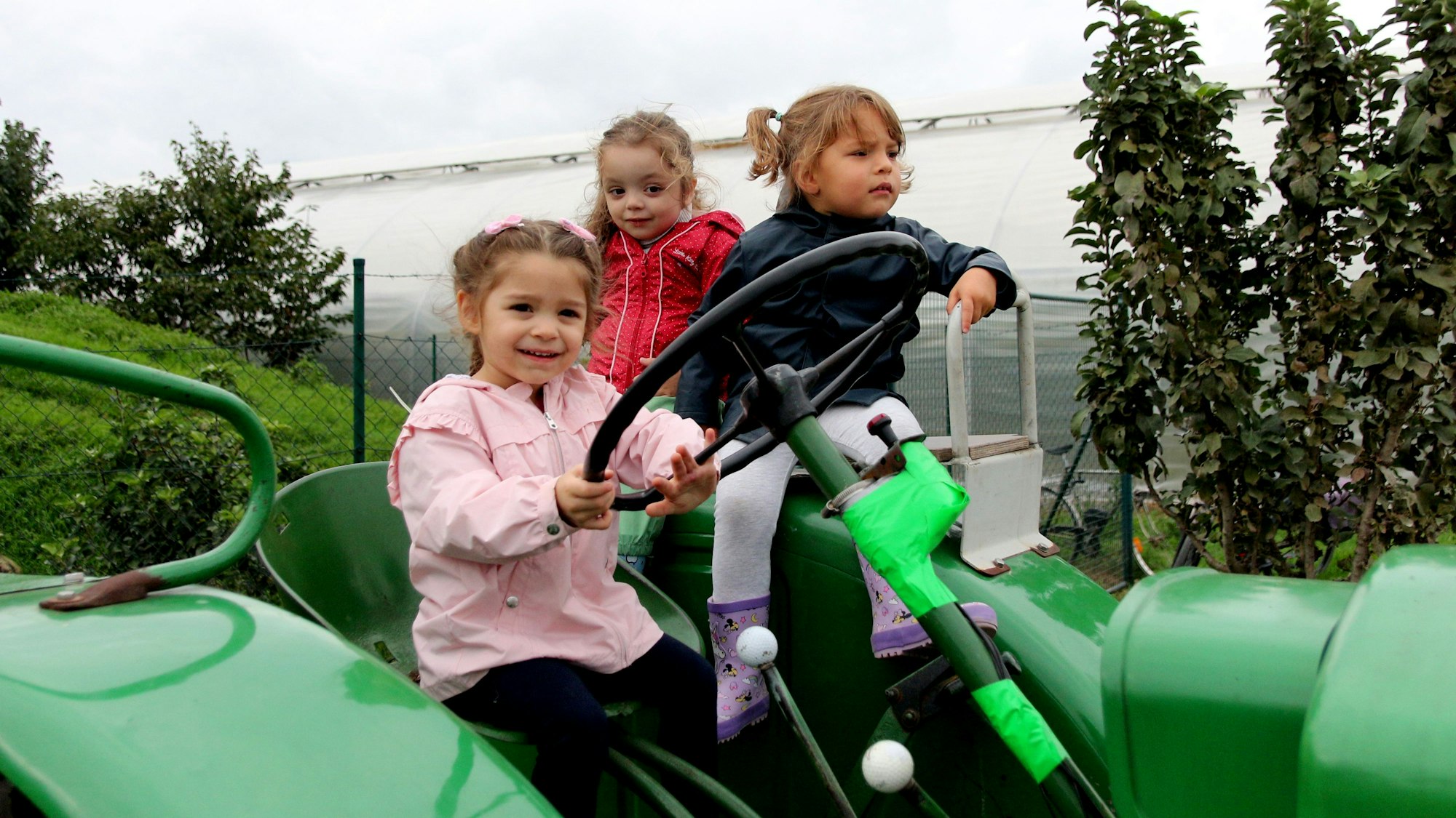 Drei Mädchen sitzen auf einem Trecker, eines umfasst sichtbar stolz den riesigen Lenker des Vehikels.