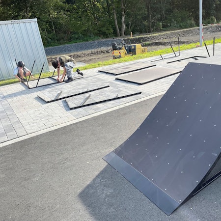 Die Rampe eines mobilen Skateparks ist zu sehen. Im Hintergrund schrauben zwei Jugendliche weitere Elemente zusammen.