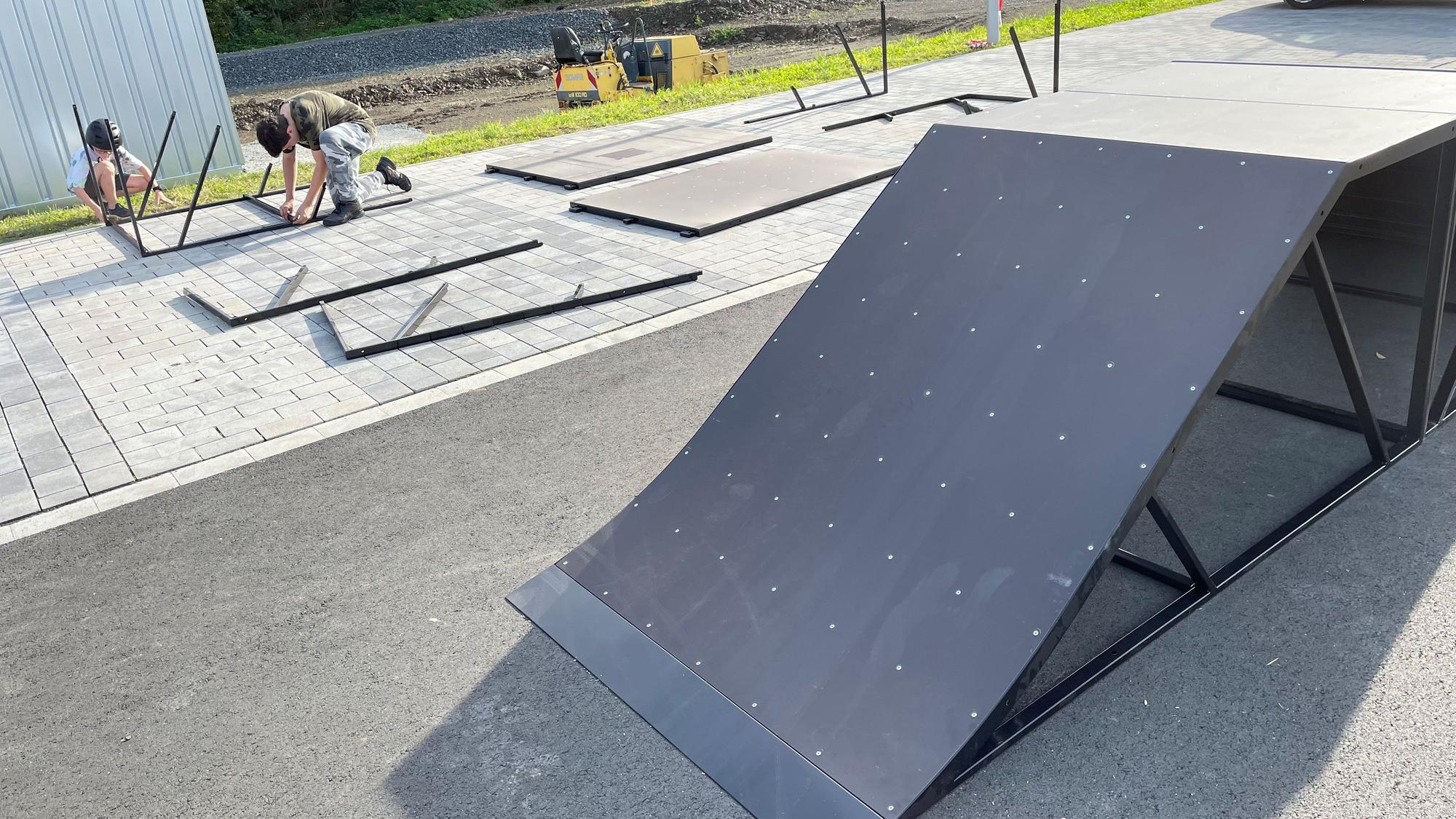 Die Rampe eines mobilen Skateparks ist zu sehen. Im Hintergrund schrauben zwei Jugendliche weitere Elemente zusammen.