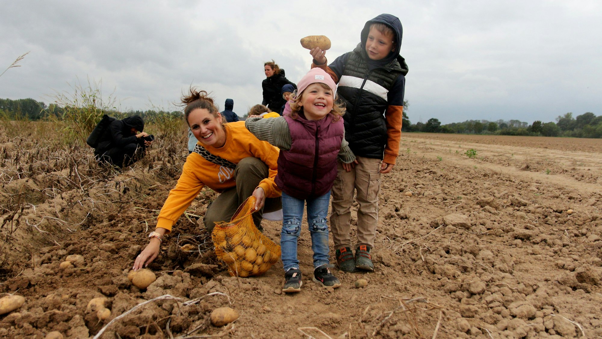 Eine Mutter kniet auf einem Acker und sammelt Kartoffeln ein. Ihre beiden Kindern grinsen in die Kamera.