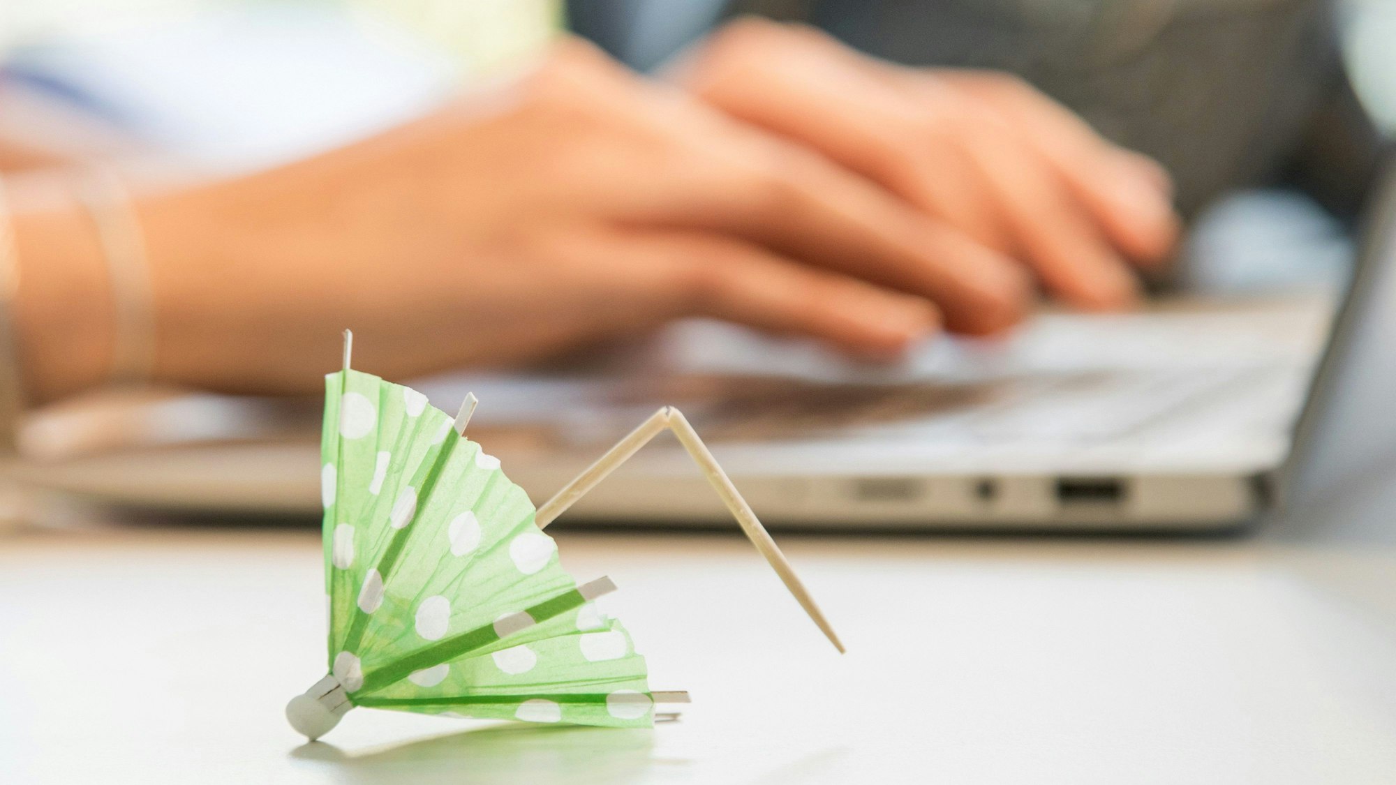 Ein umgeknicktes Papierschirmchen liegt vor einem Laptop auf dem zwei Hande tippen.