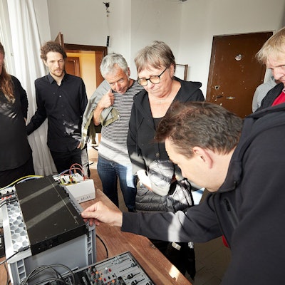 Eine Gruppe Menschen steht um einen Tisch, auf dem elektronische Geräte aufgebaut sind. Ein Mann bedient einen Regler.