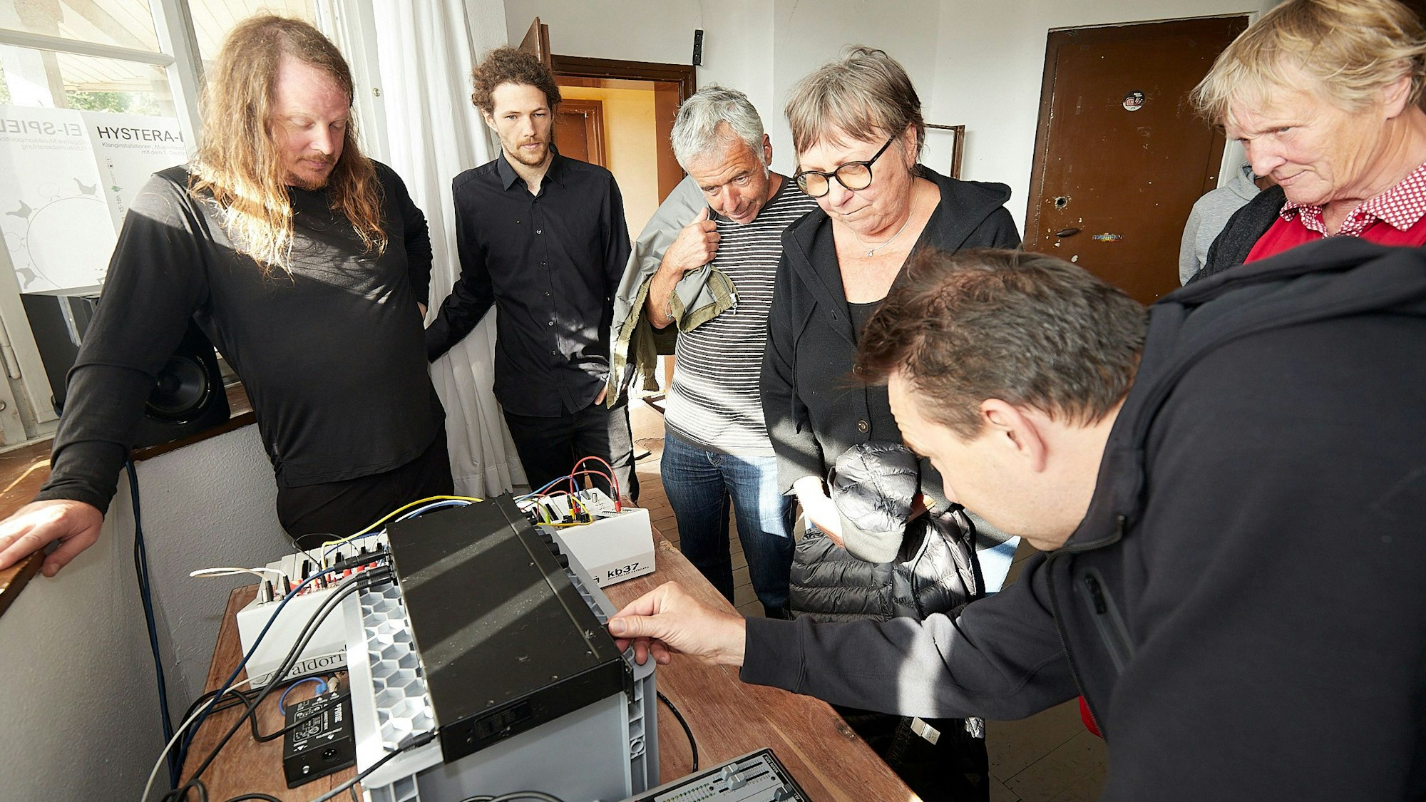 Eine Gruppe Menschen steht um einen Tisch, auf dem elektronische Geräte aufgebaut sind. Ein Mann bedient einen Regler.