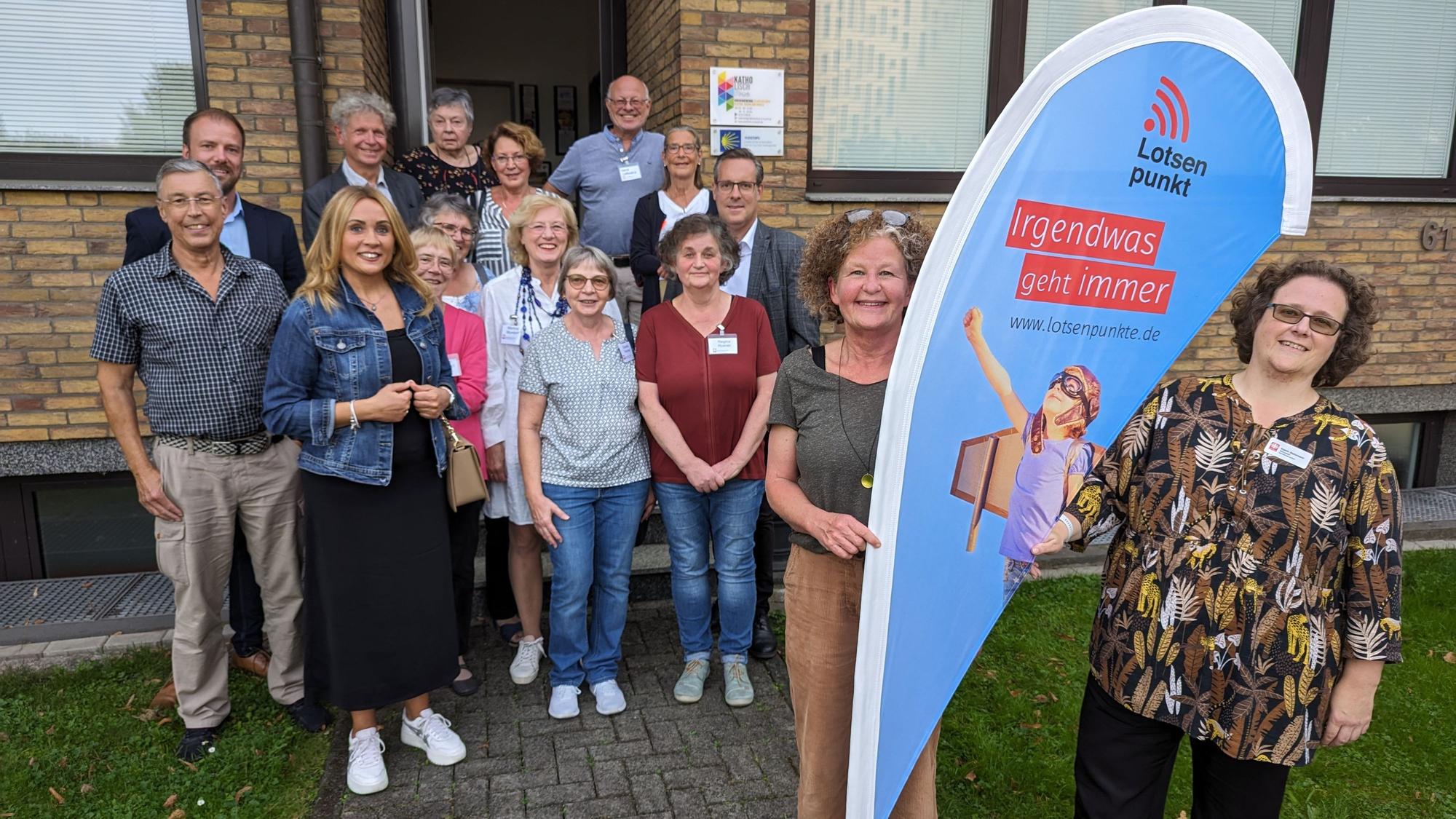Eine Gruppe von Frauen und Männern vor einem Haus, im Fordergrund zwei Frauen an einer Flagge mit der Aufschrift Lotsenpunkt.
