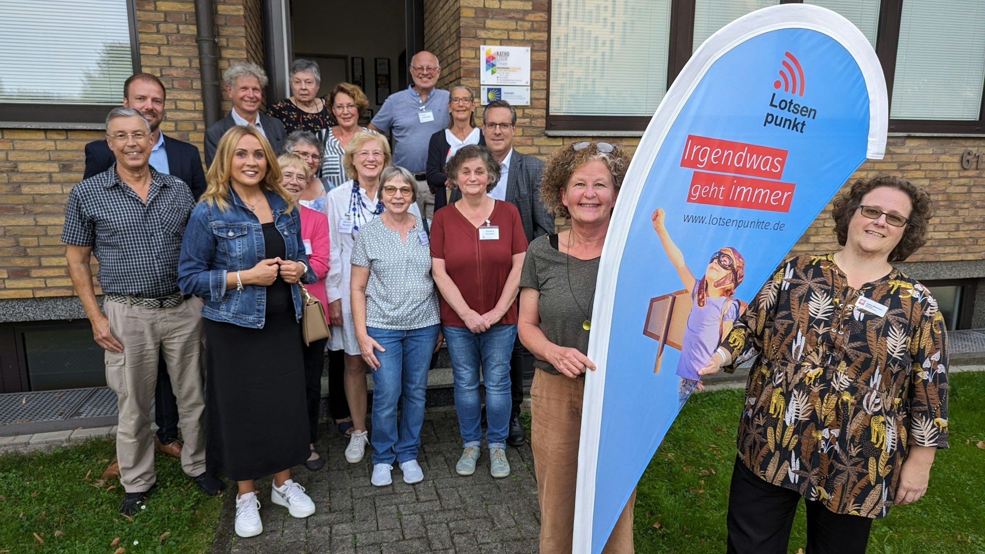 Eine Gruppe von Frauen und Männern vor einem Haus, im Fordergrund zwei Frauen an einer Flagge mit der Aufschrift Lotsenpunkt.